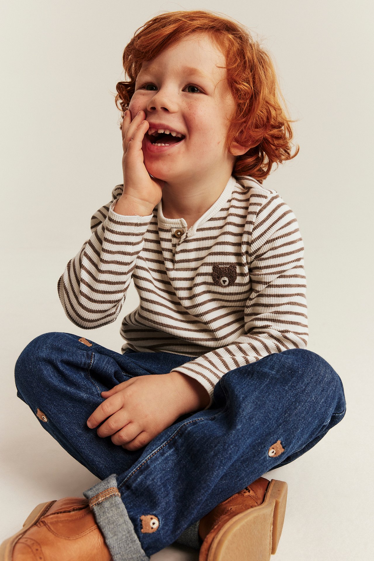 Front view of a kid wearing a ribbed striped henley top with bear embroidery and blue jeans with bear embroidery.