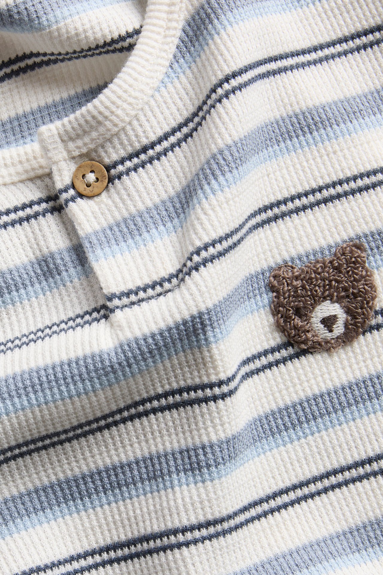 Close-up of a baby's cream and blue striped waffle knit top with a bear appliqué and button.