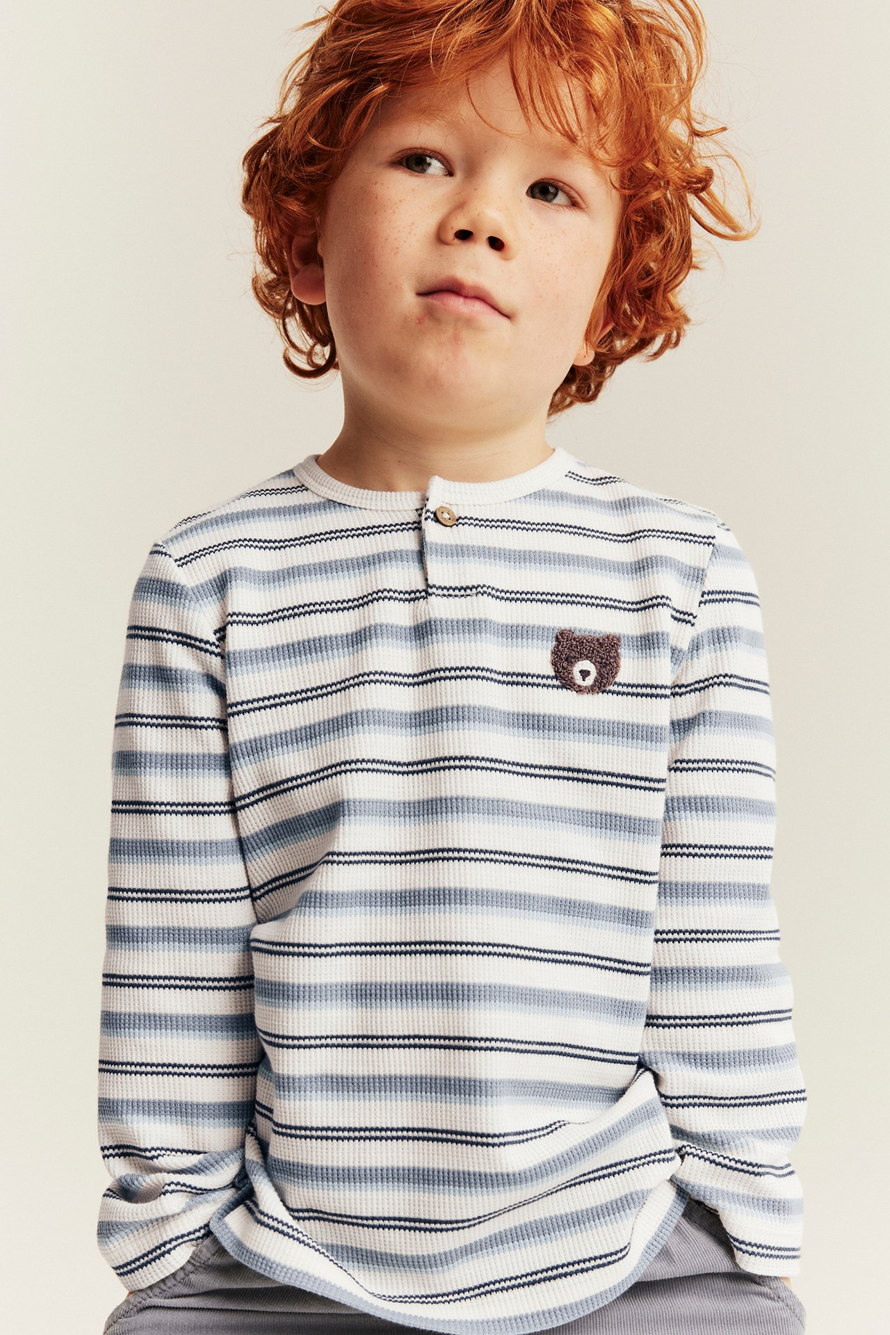 Front view of a kid's striped ribbed long-sleeve Henley shirt with a teddy bear embroidery, paired with gray pants.