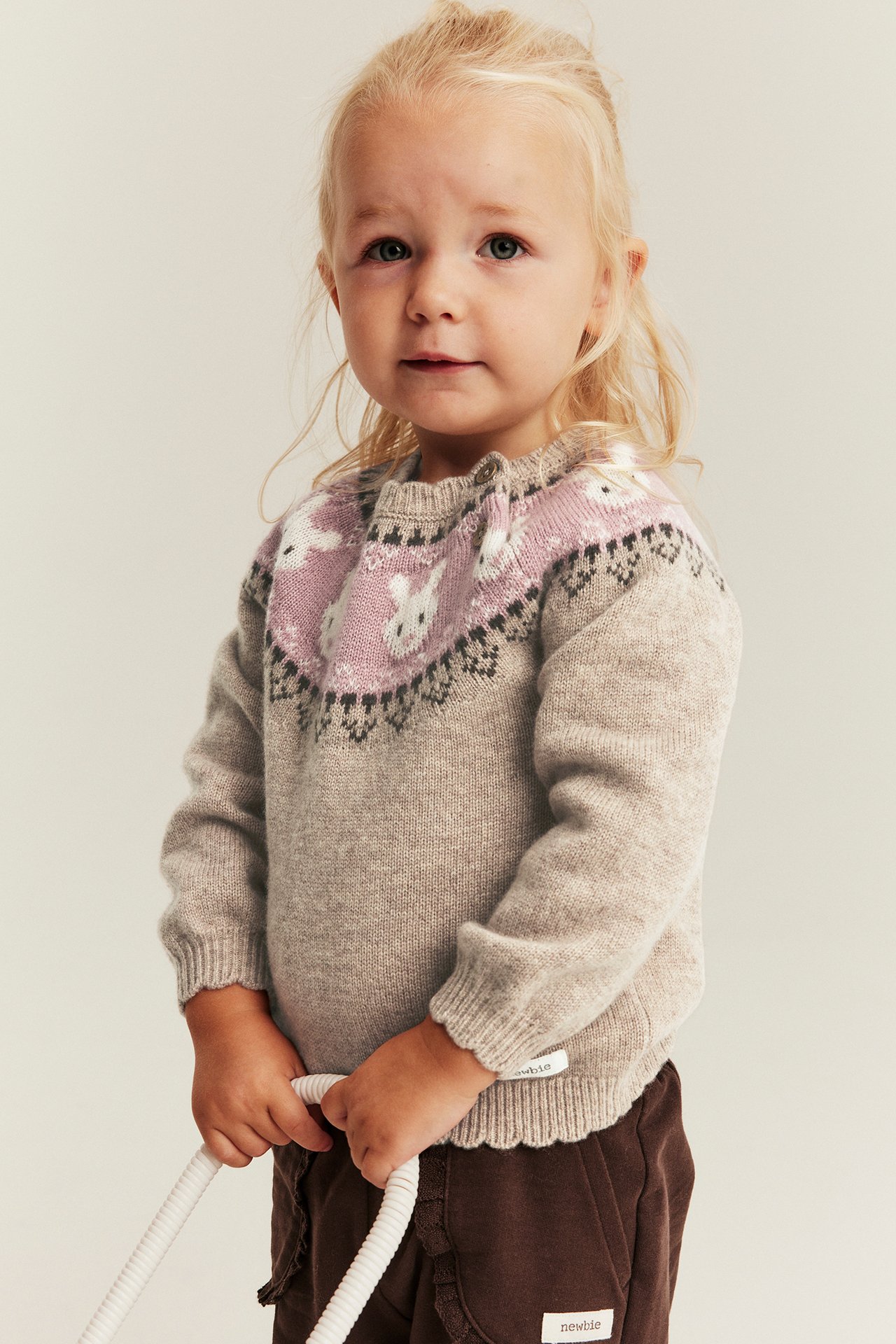 Vorderansicht: Kind in beigem Strickpullover mit rosa Hasenpasse, dunkelbrauner Hose, hält weißes Seil.