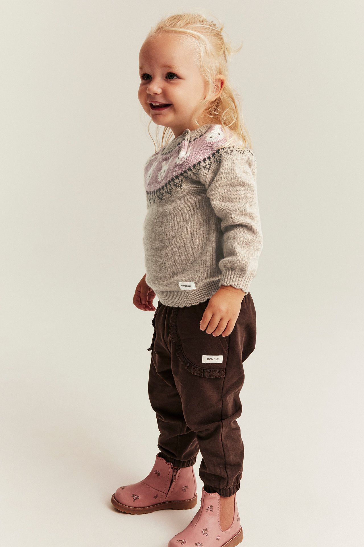 Seitenansicht eines Kindes in beigem gemustertem Strickpullover, dunkelbrauner Hose und rosa Blumen-Stiefeletten.