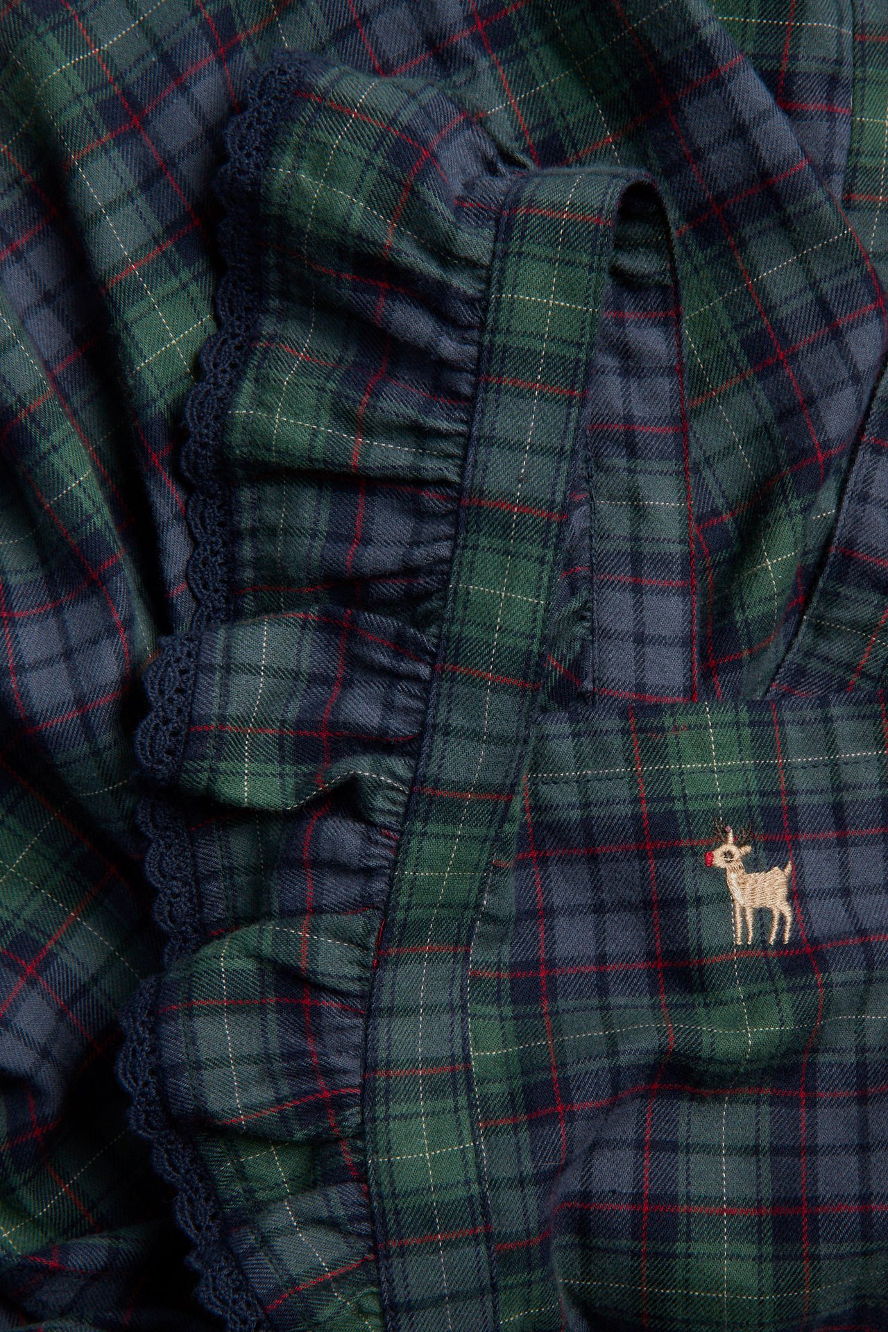 Close-up of green and blue plaid overall dress with ruffled strap, lace trim, deer embroidery.