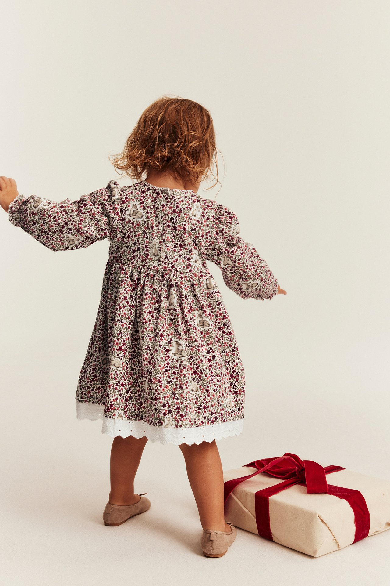 Rückansicht eines Kindes in rotem Blumenkleid mit Spitzenbesatz und beigen flachen Schuhen.