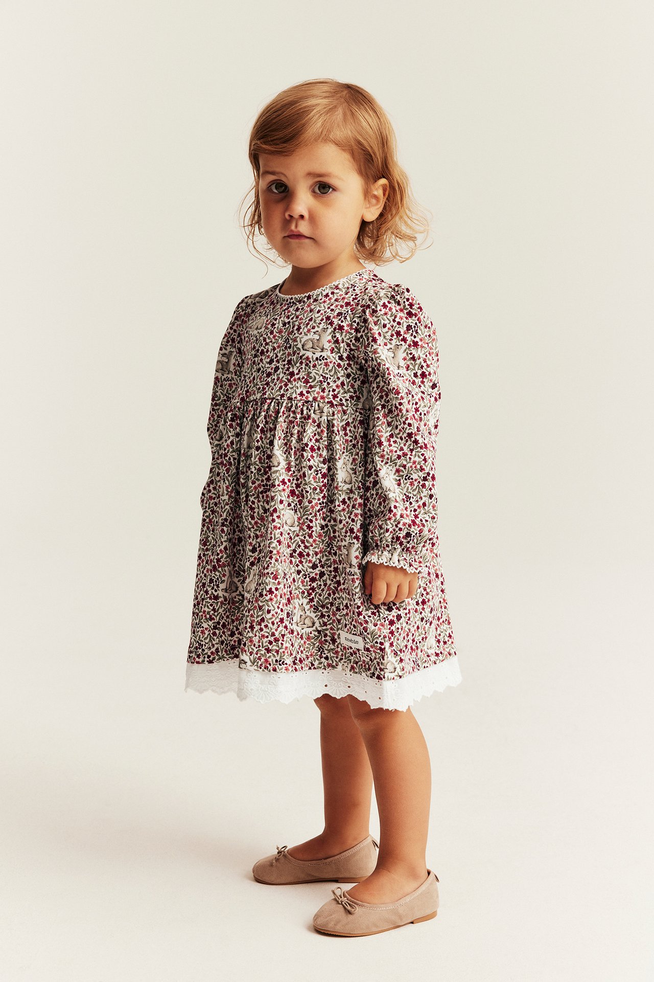 Vorderansicht eines Kinderkleides mit langen Ärmeln, Blumen- und Hasenmuster, weißer Spitze und beigen Ballerinas.