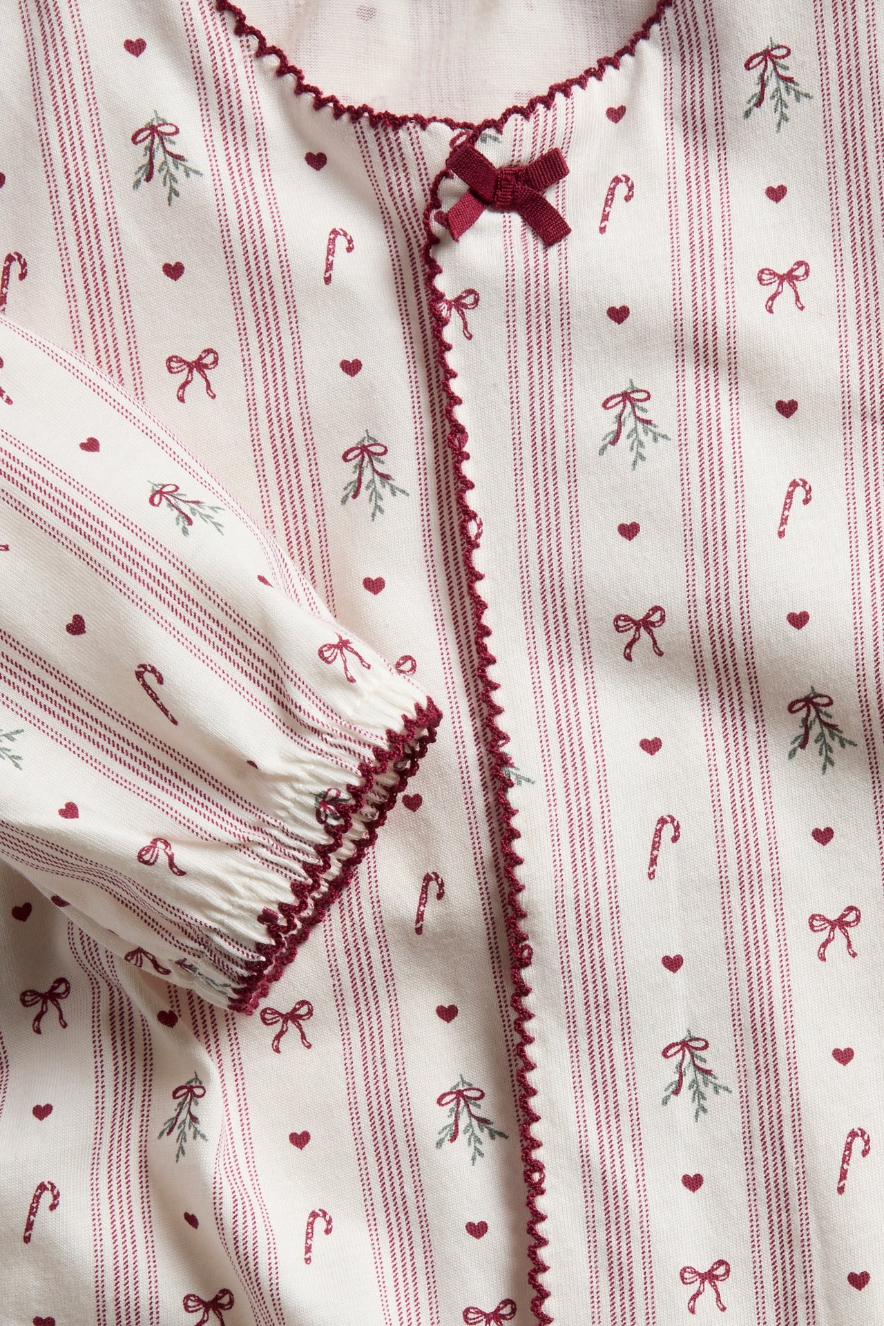 Close-up of a cream pajama top with red stripes, holiday patterns, red picot trim, and a red bow.