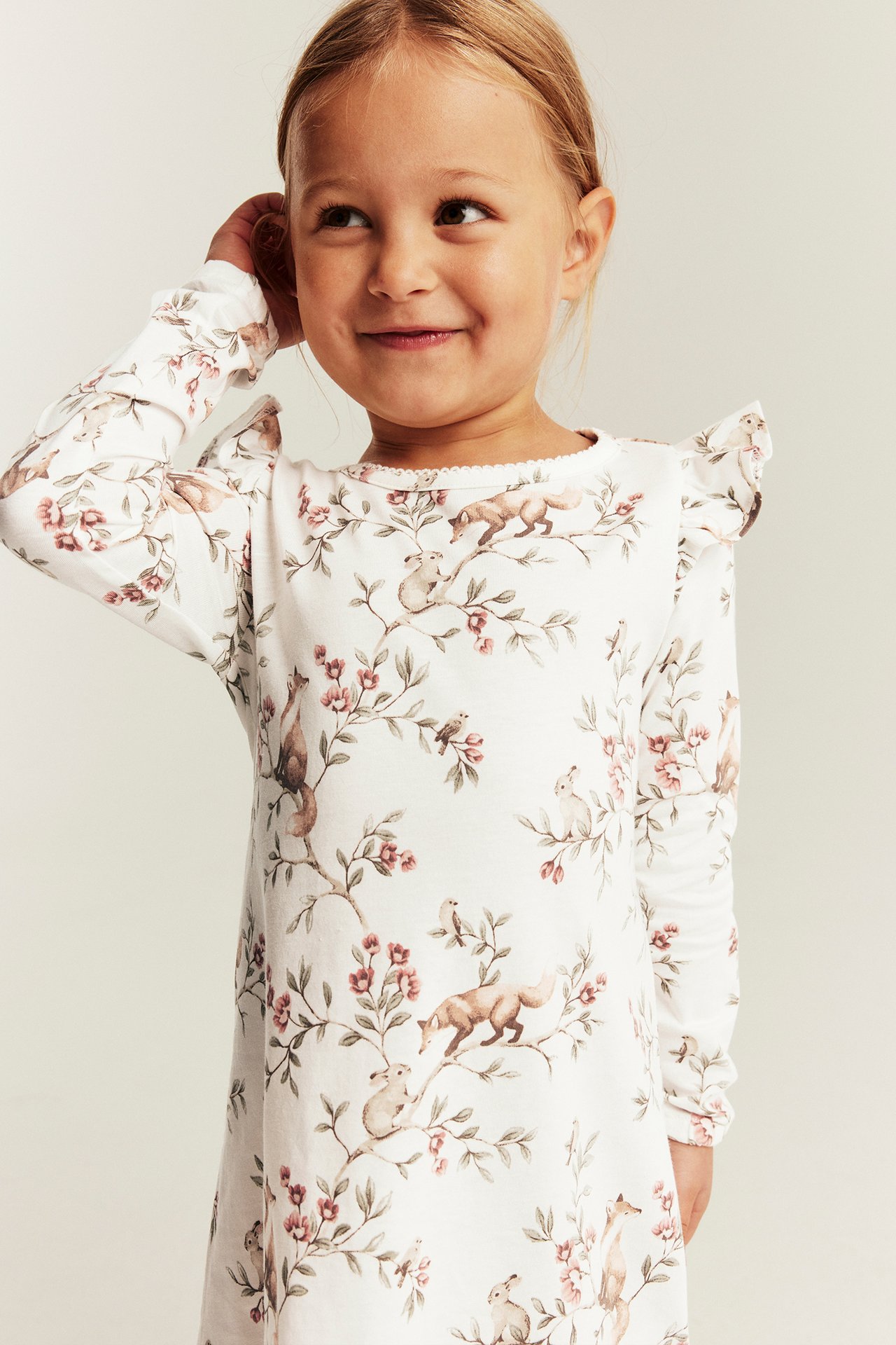 Vorderansicht: Kind in weißem Langarmkleid mit Schulterrüschen und Waldtiermuster.