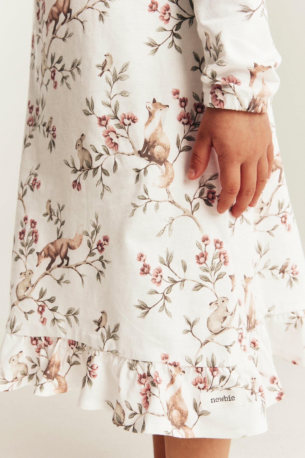 Nahaufnahme eines weißen langärmeligen Kinderkleides mit Waldtier- und rosa Blumenmuster und Rüschensaum.