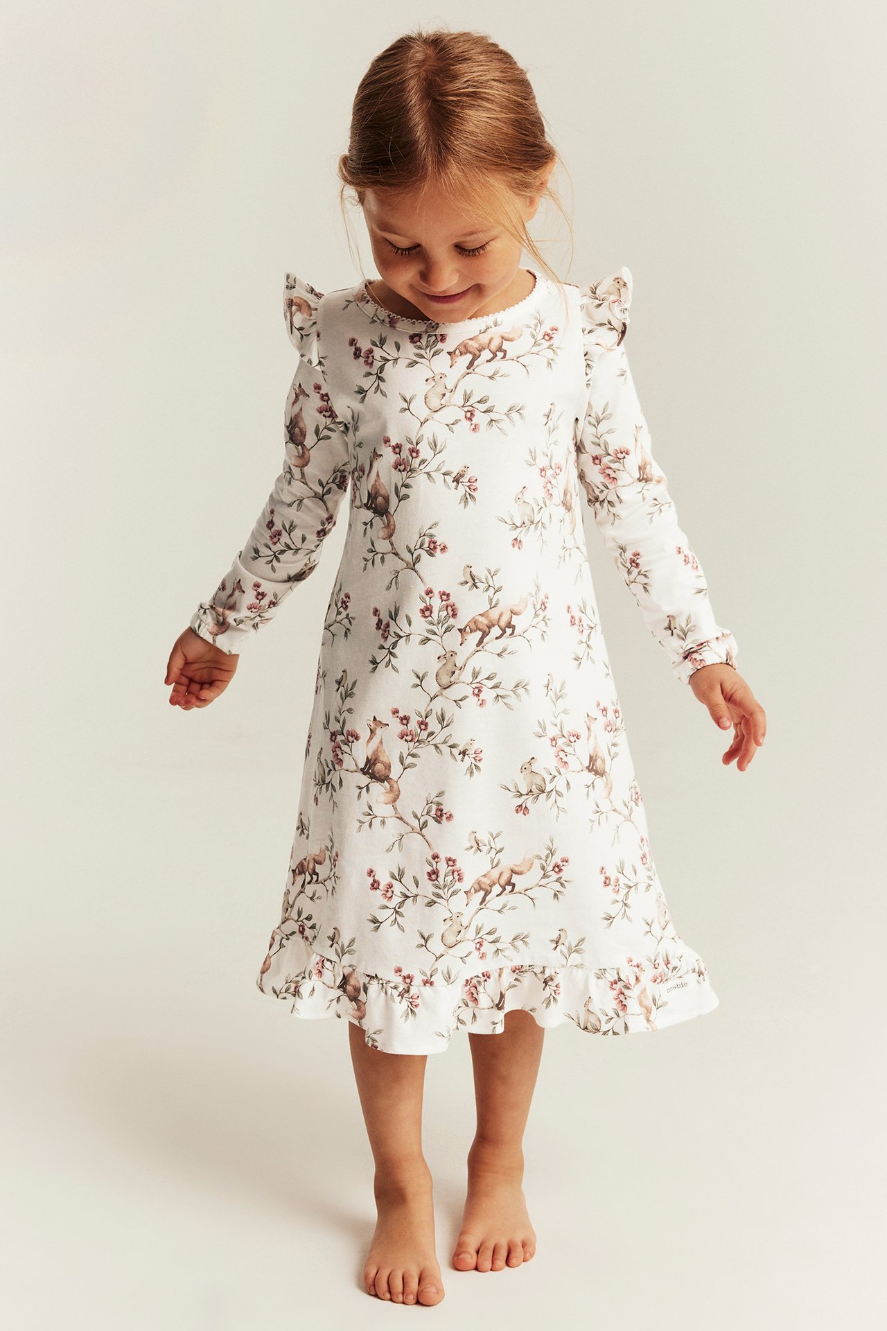 Vorderansicht eines Kindes in einem weißen langärmeligen Rüschenkleid mit Tier- und Blumenmuster.