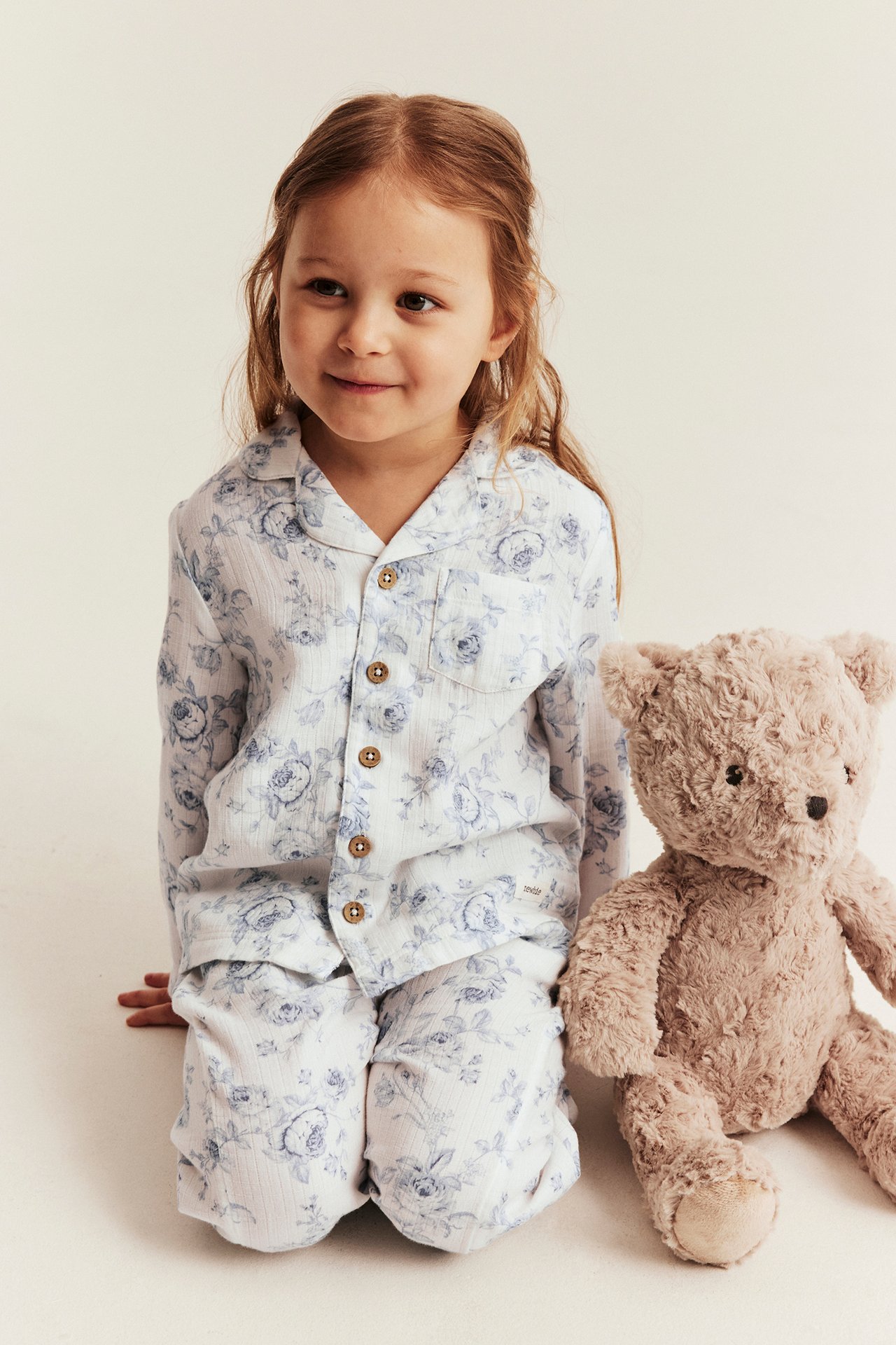 Front view of a kid in white cotton floral pajamas with buttons, beside a brown teddy bear.
