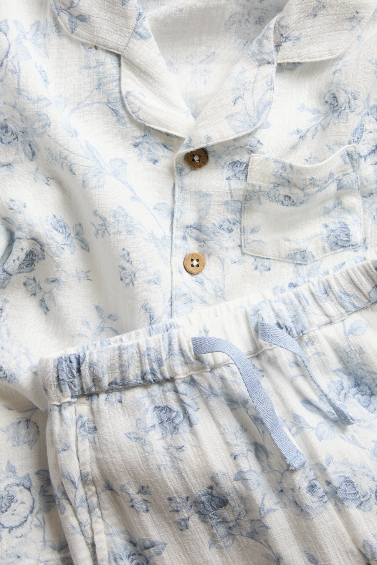 Close-up of white cotton two-piece pyjamas with light blue floral print, collared shirt, and drawstring waist pants.