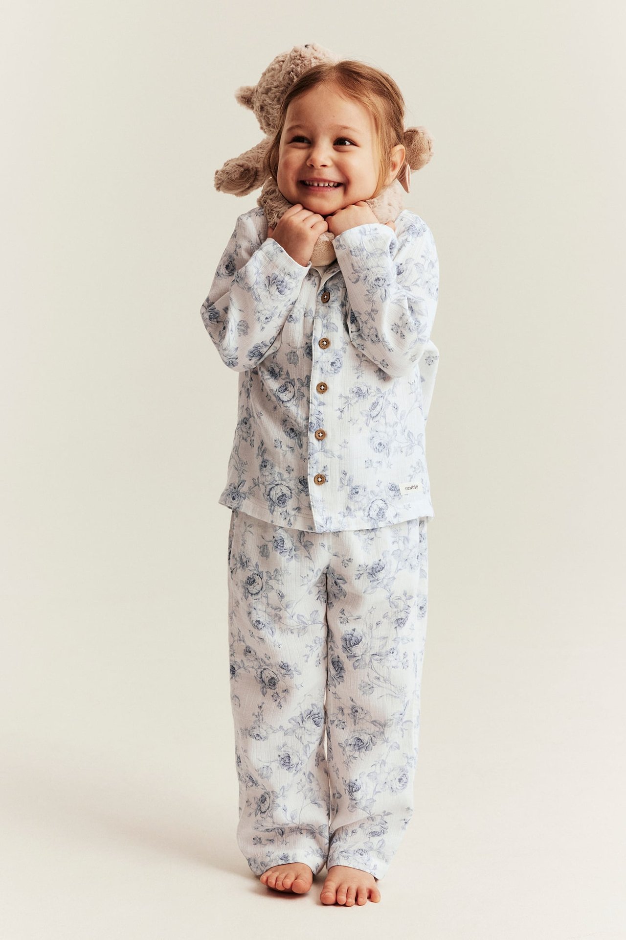 Vorderansicht: lächelndes Kind trägt weißen Baumwollpyjama mit Blumenmuster, Hemd mit Knöpfen, Hose, hält braunen Teddybären.