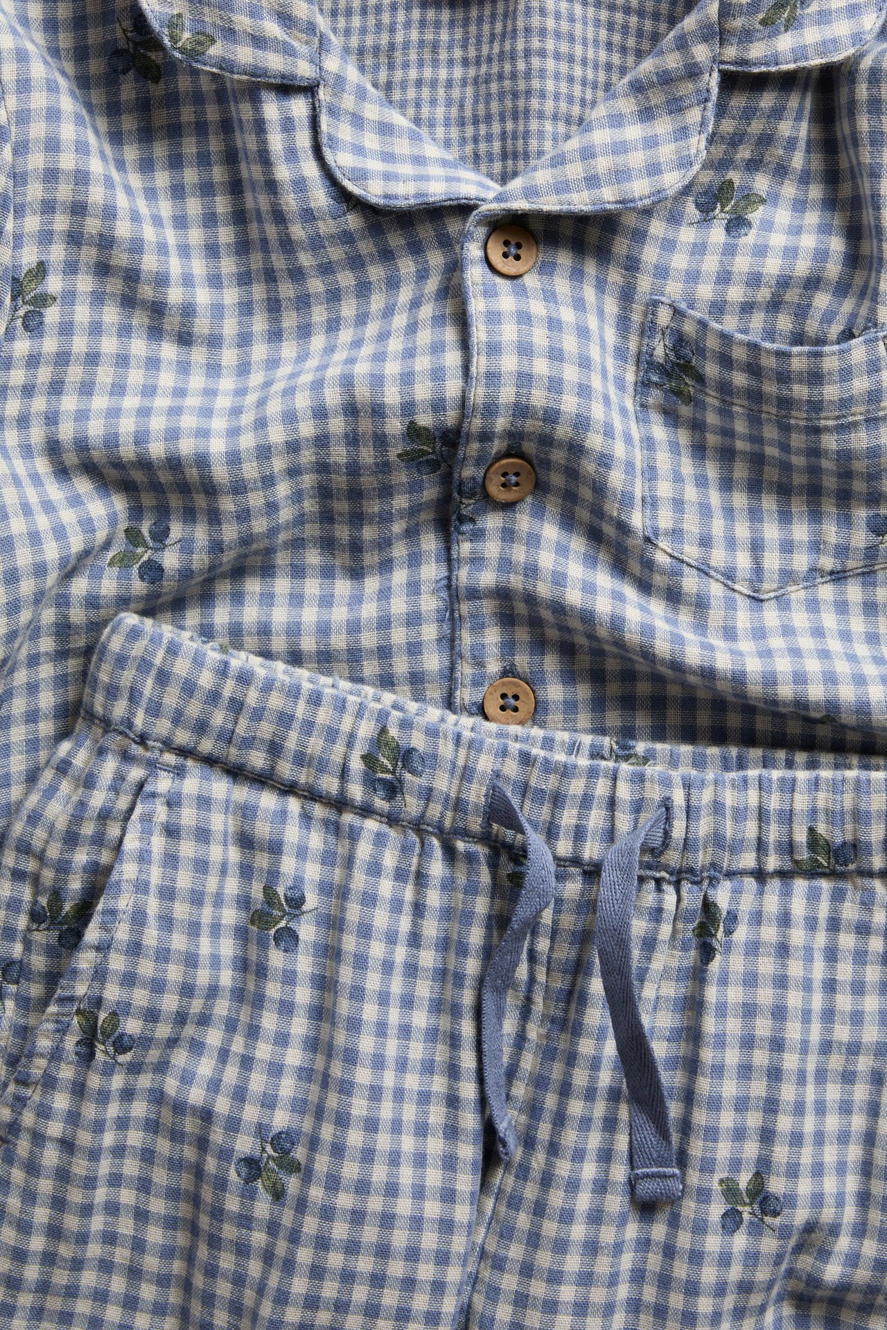 Close-up of blue and white gingham cotton pajamas with fruit pattern. Collared shirt, button front, pants with drawstring.