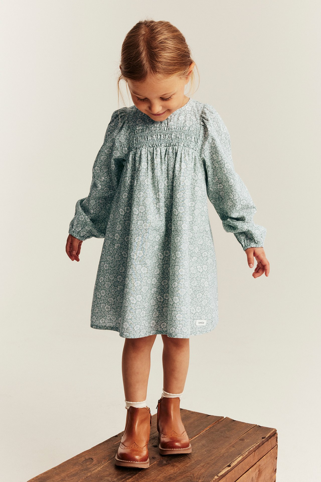 Vorderansicht eines Kindes in hellblauem geblümten Smockkleid, braunen Stiefeletten und weißen Socken.