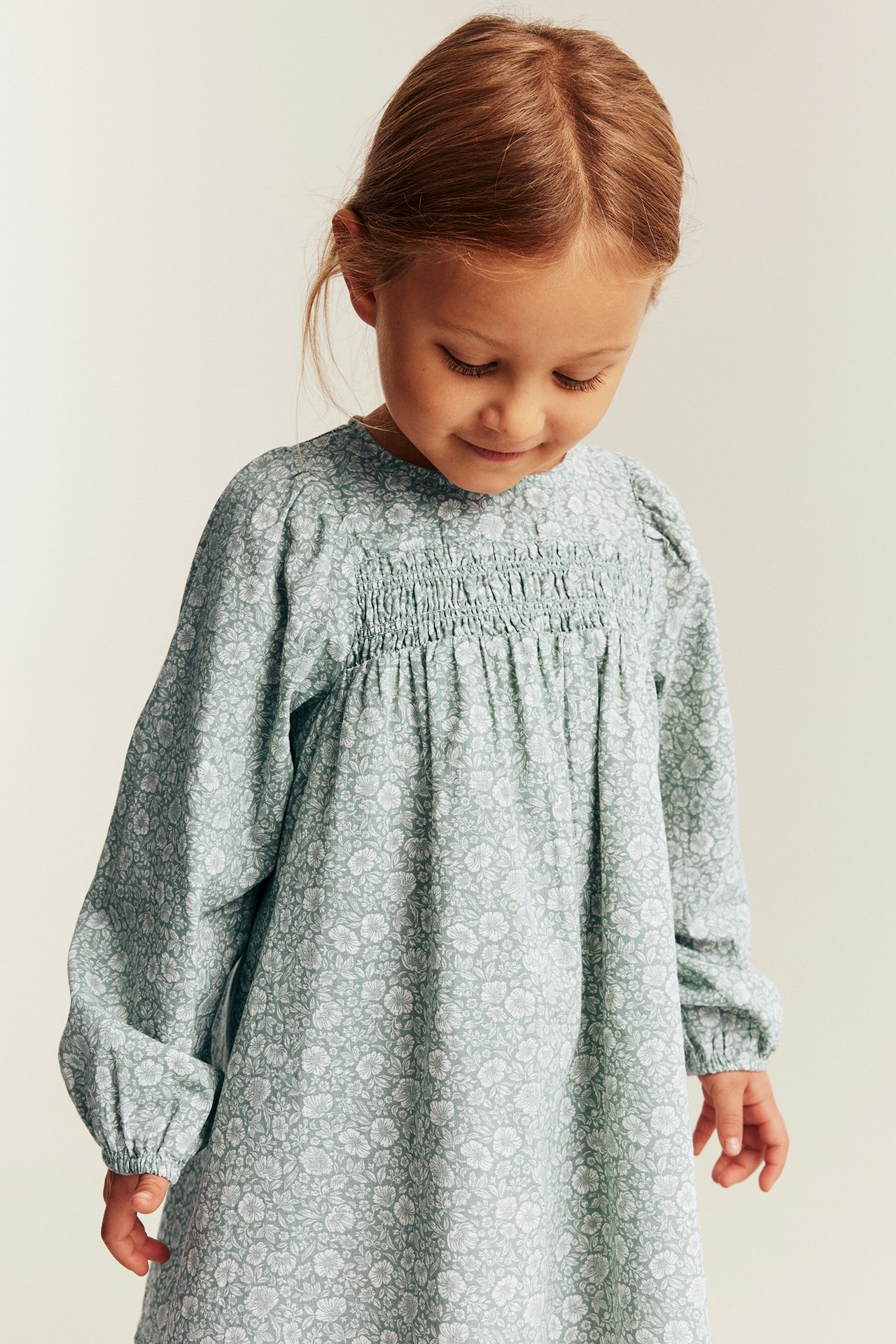 Ein hellgrünes geblümtes langärmeliges Kinderkleid mit gesmoktem Oberteil, Vorderansicht.