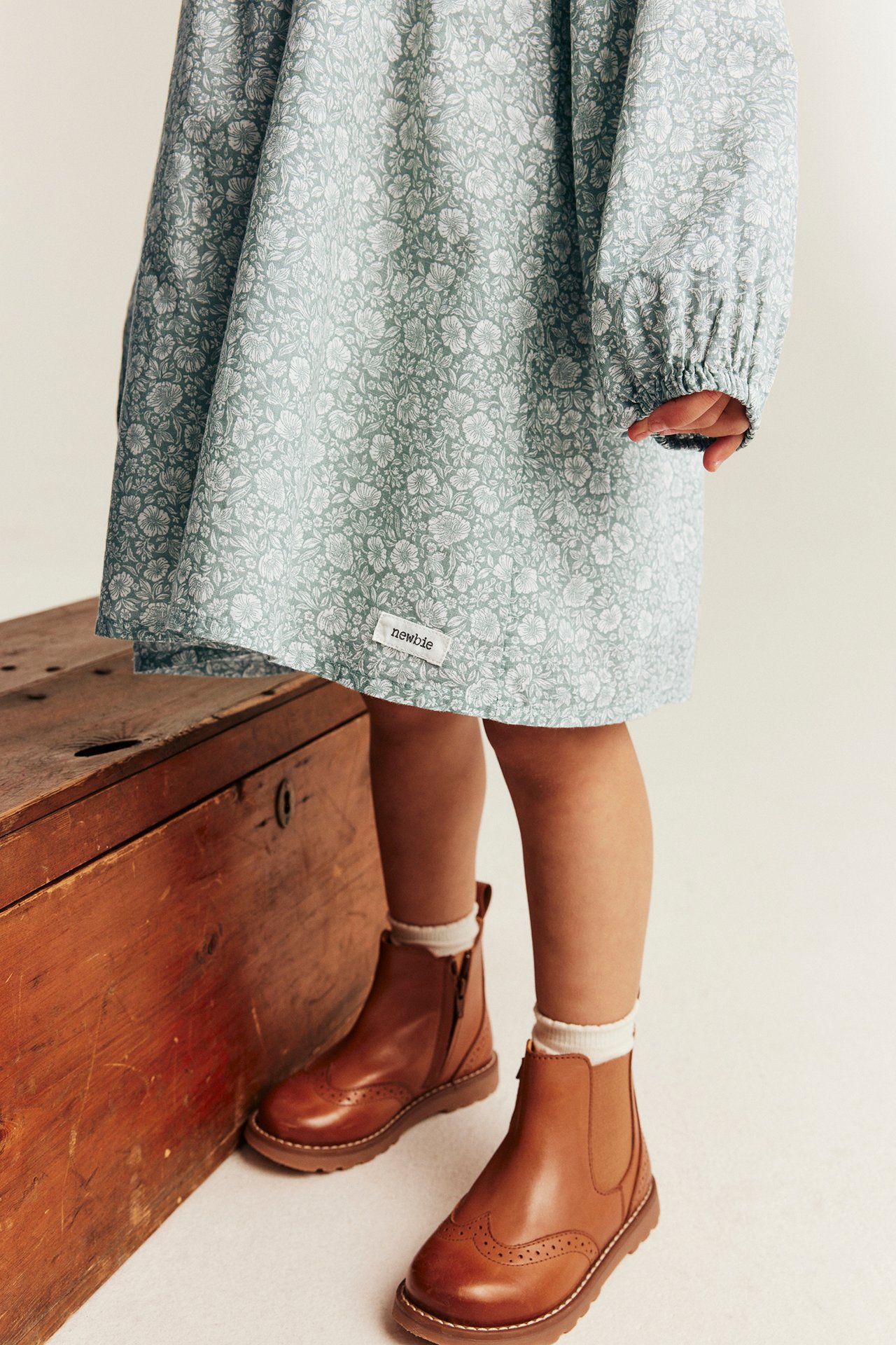 Vorderansicht: Kind in hellgrünem Blumenkleid mit langen Ärmeln und braunen Leder-Brogue-Stiefeln.