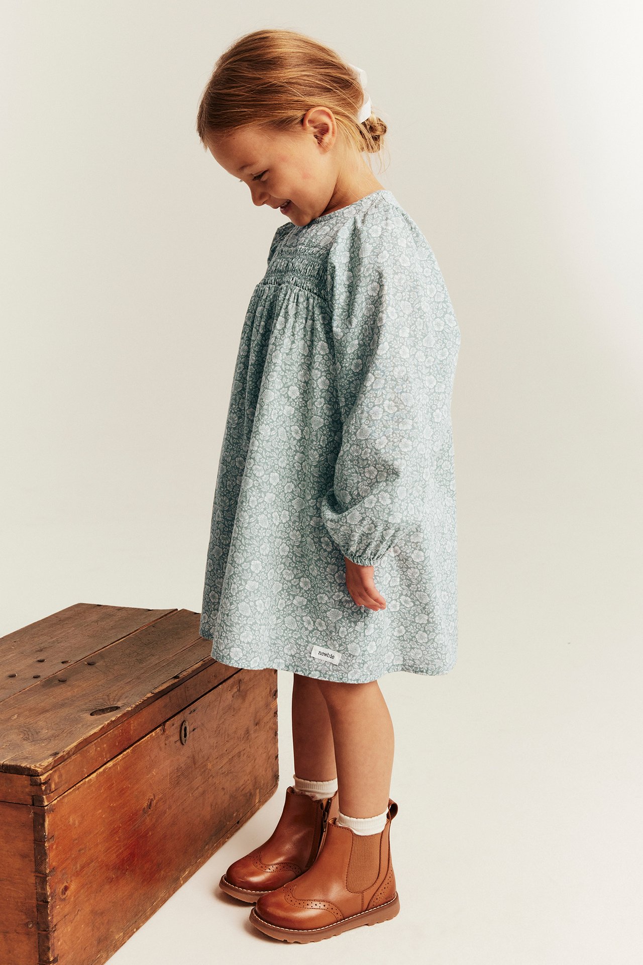 Seitenansicht eines Kindes in hellgrünem floralem Langarm-Smockkleid, weißen Socken und braunen Lederstiefeln.