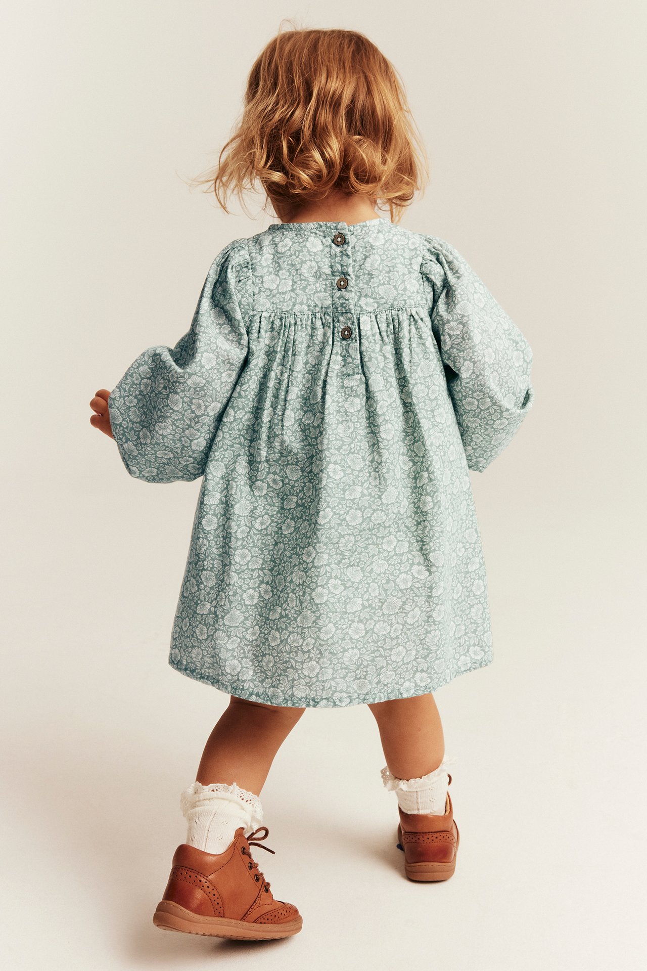 Rückenansicht: Baby in hellgrünem Blumenkleid mit Knöpfen, weißen Socken und braunen Schuhen.