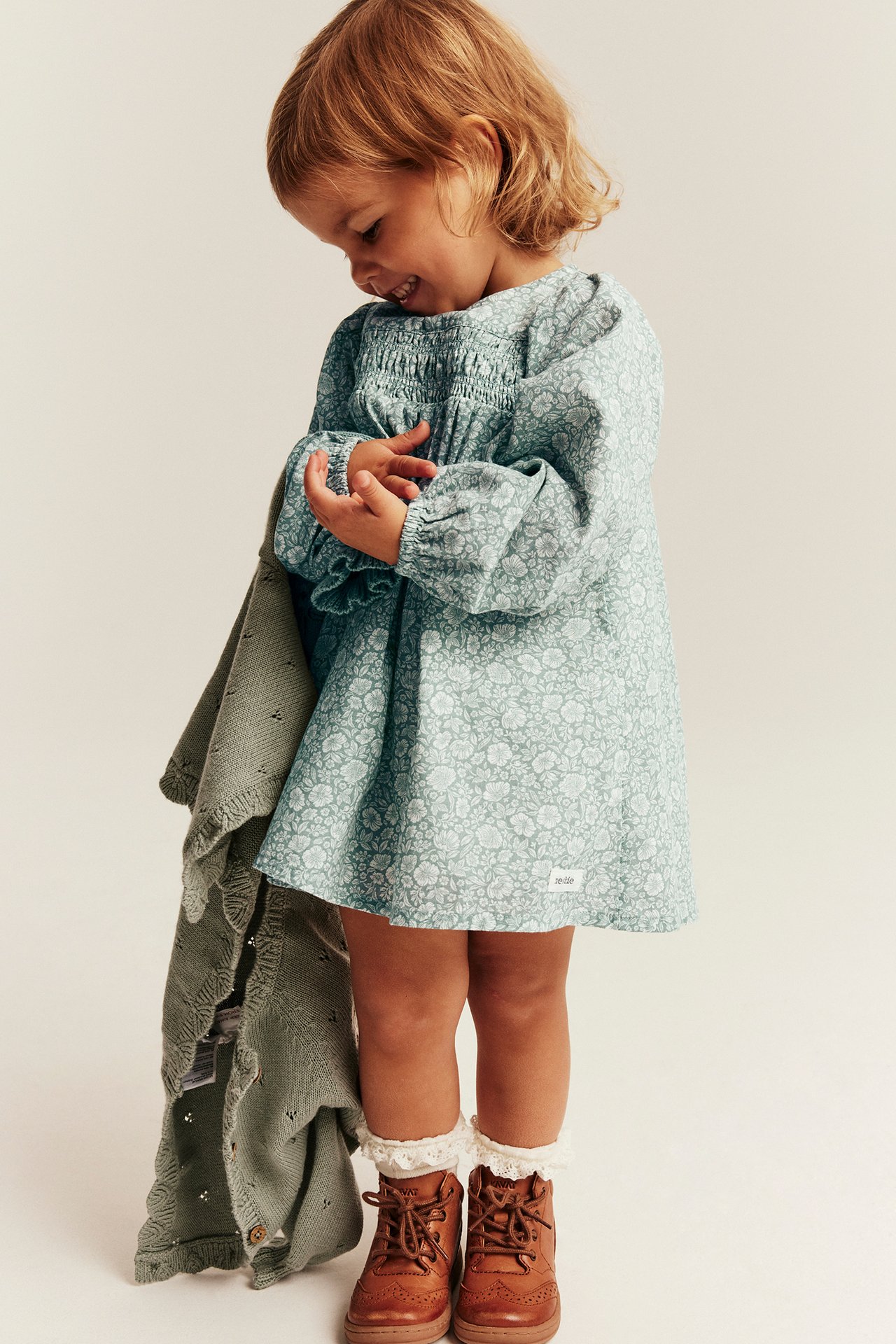 Kid wears a light blue floral smocked dress, holds a green knitted cardigan, brown shoes.