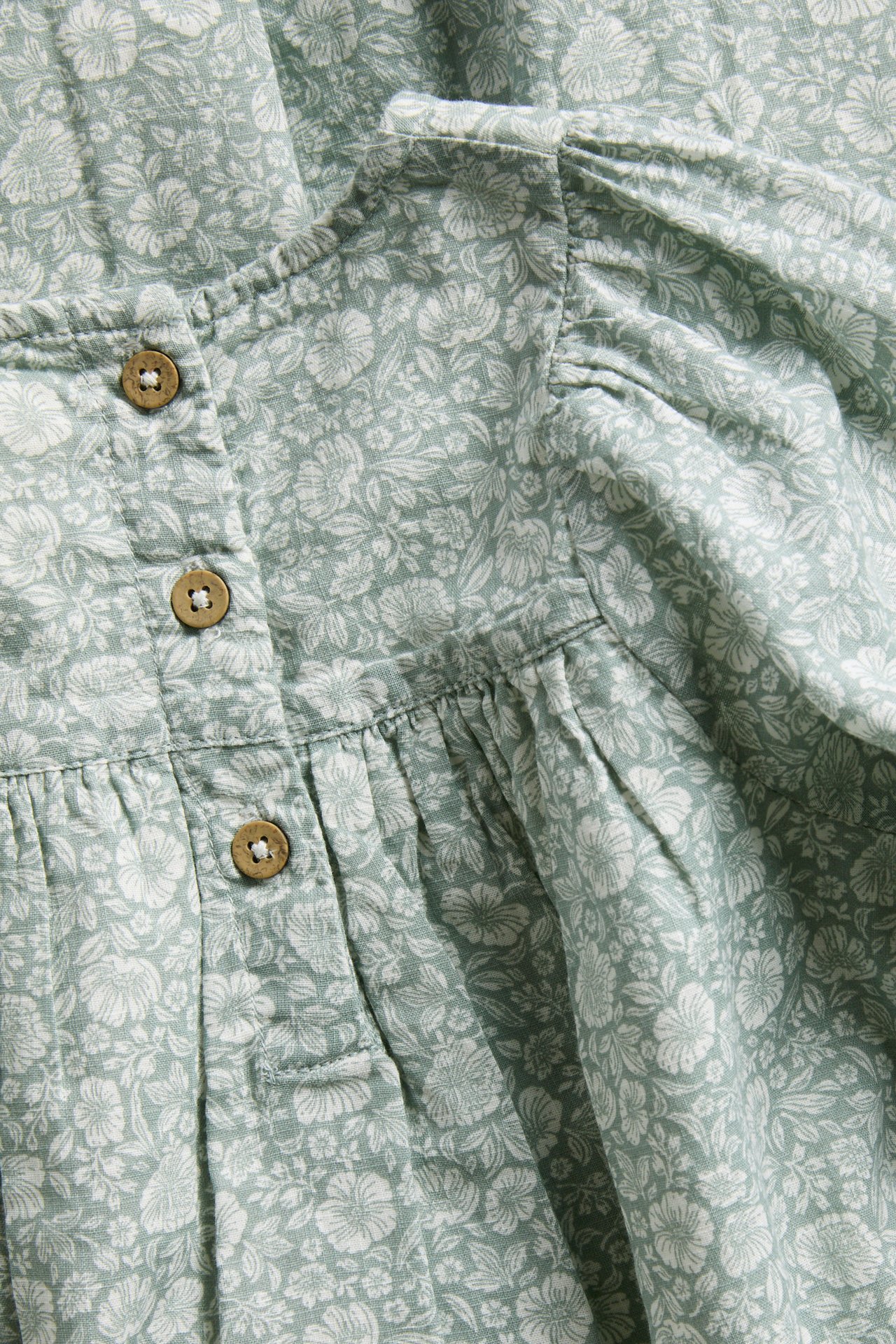 Close-up of a light green floral print garment with three brown buttons and gathered fabric.
