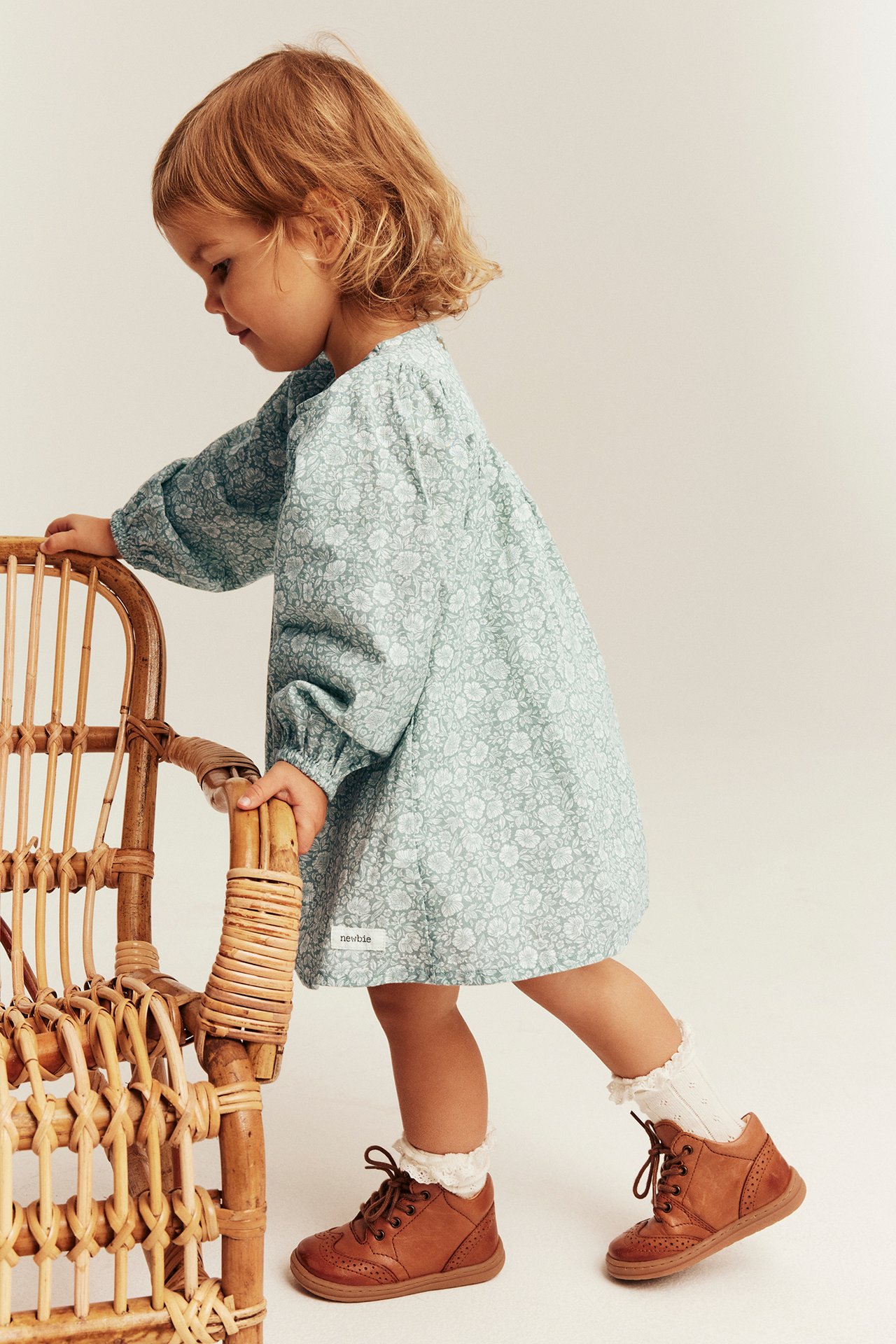 Side view: kid in light blue floral long-sleeved dress, white frill socks, and brown shoes.