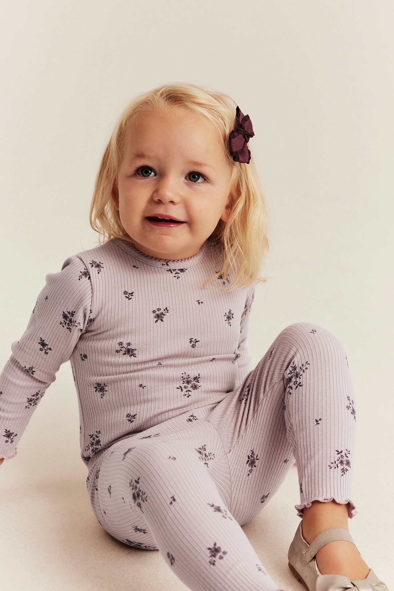 Vorderansicht von Kinder-Loungewear-Set in Helllila gerippt-geblümt, dunkler Schleife und grauen Schuhen.