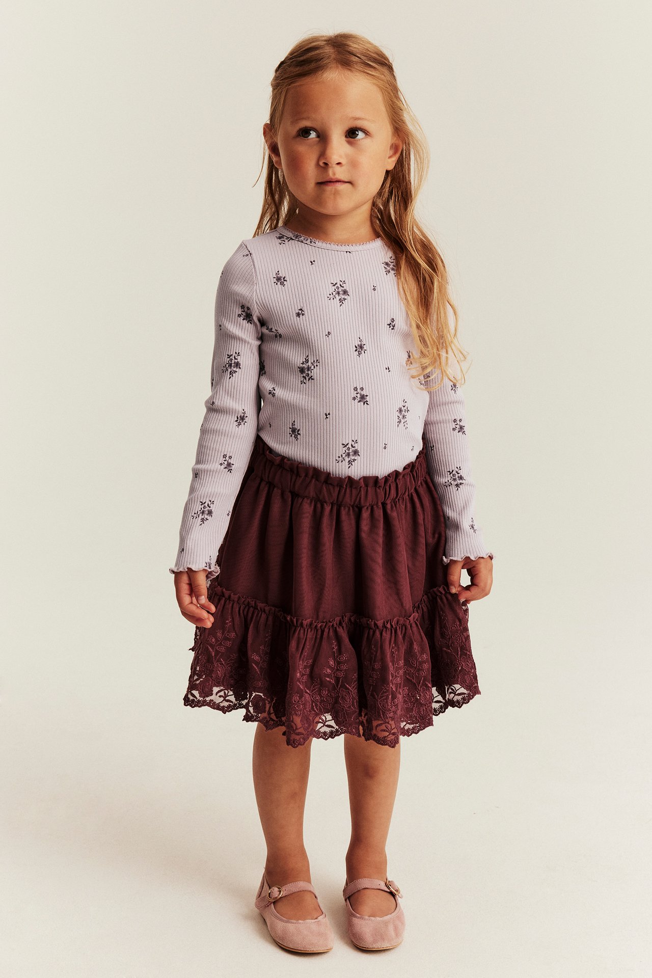Front view: kid in light purple floral long-sleeved top, maroon tiered lace skirt, and pink Mary Jane shoes.