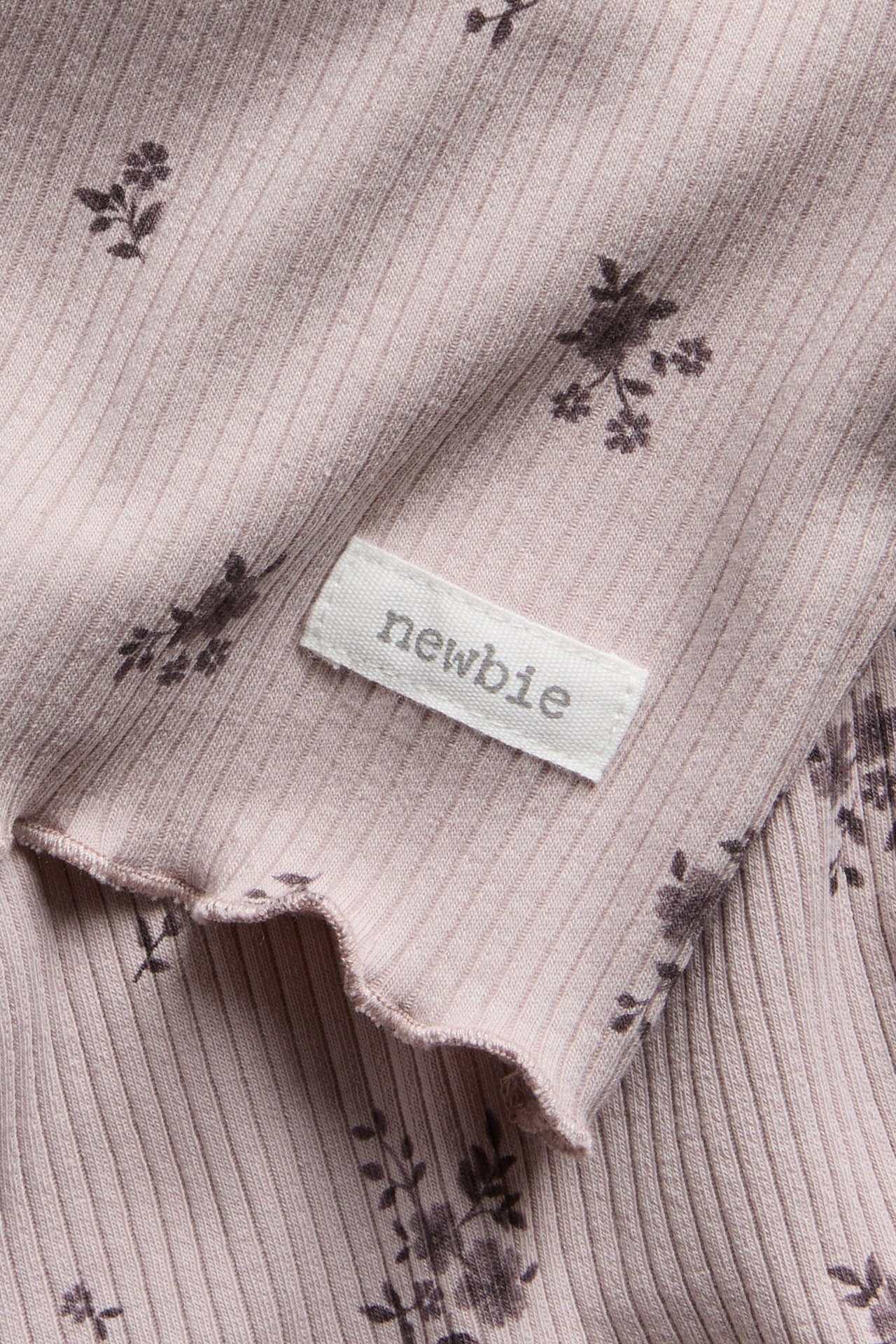 Close-up of a pink ribbed baby garment with dark floral print, lettuce edge, and a 'newbie' label.