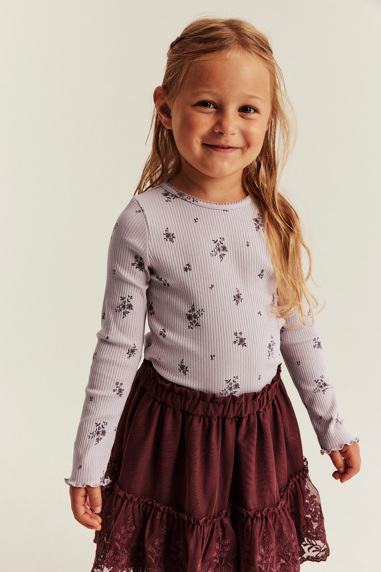 Front view of a kid wearing a lilac ribbed floral long-sleeve top and a burgundy tiered ruffled skirt.