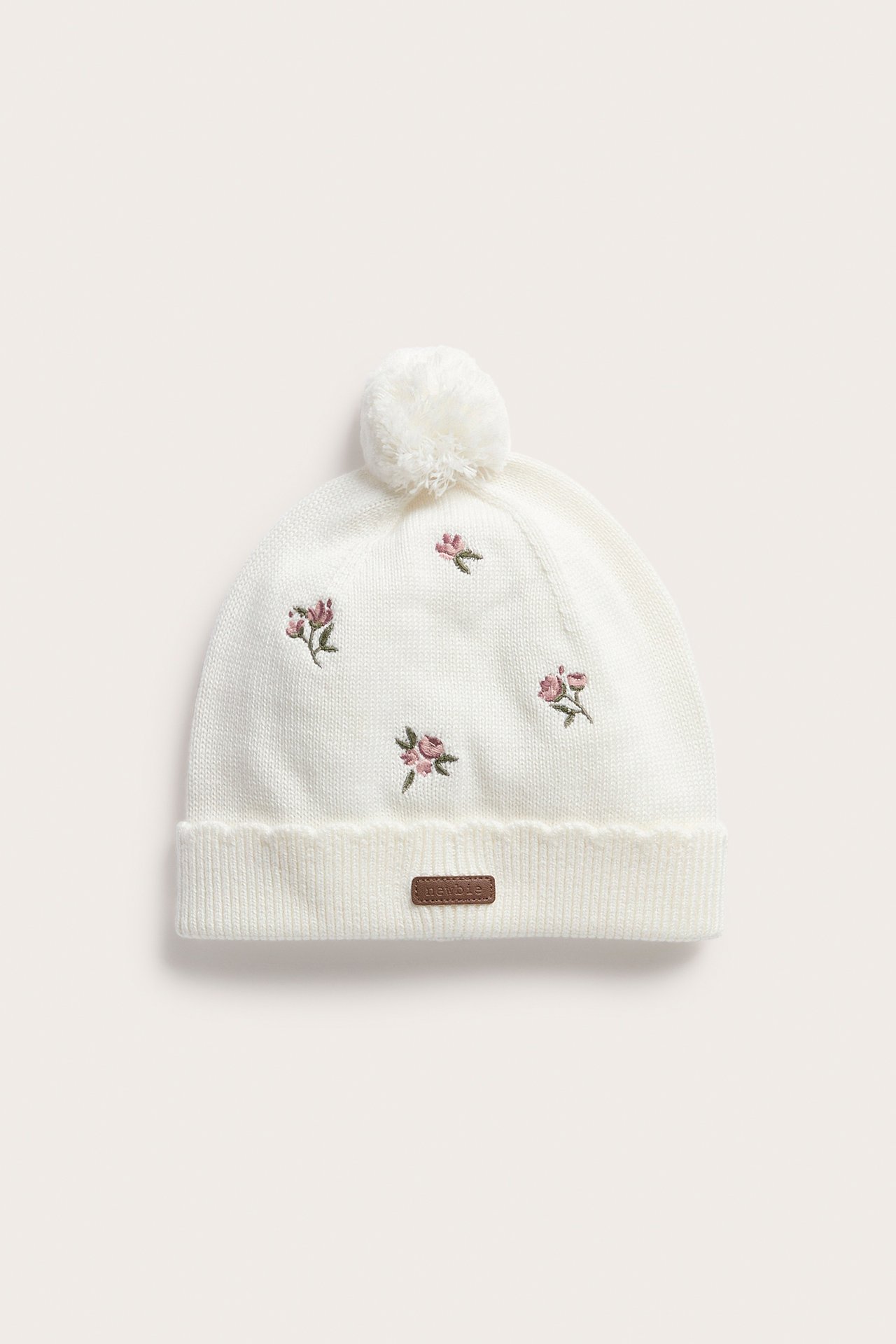 Front view of a white knitted baby beanie with a pom-pom, floral embroidery, and a brown label.