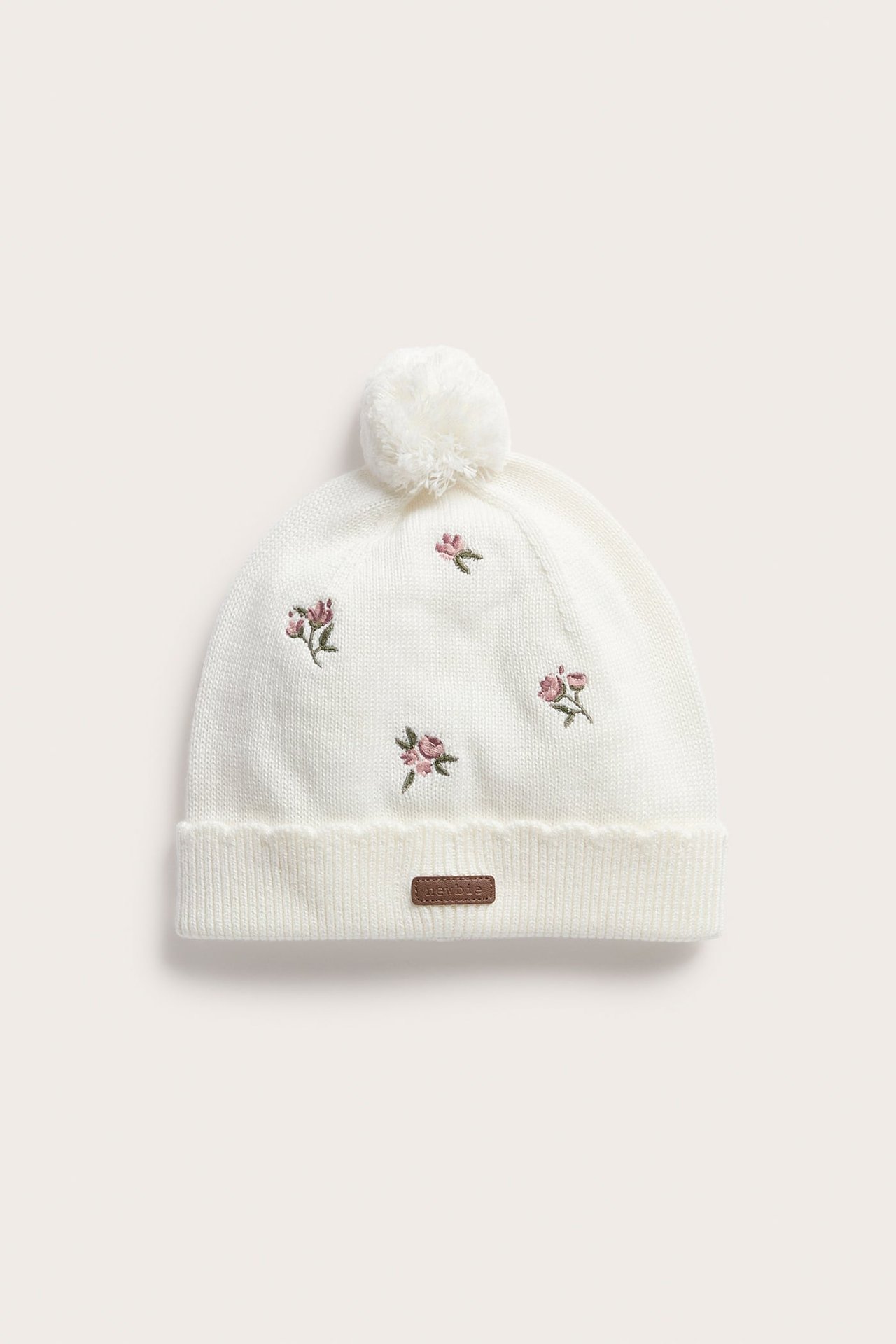 Vorderansicht einer weißen gestrickten Babymütze mit Bommel, Blumenstickerei und braunem Etikett.