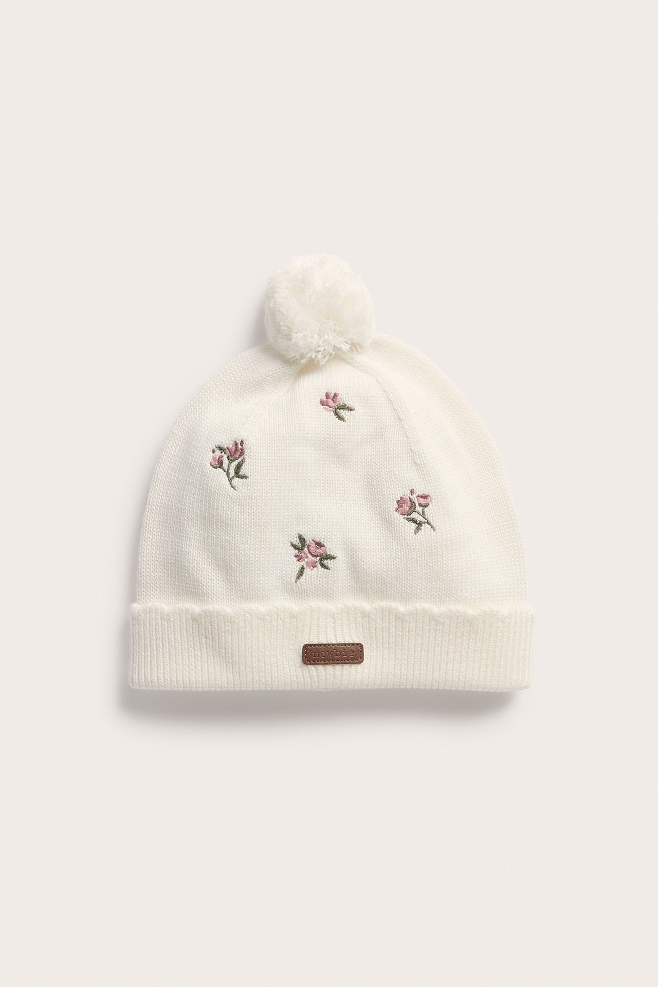 Vorderansicht: Eine weiße gestrickte Babymütze mit rosa Blumenstickerei, Pompon und braunem Etikett.
