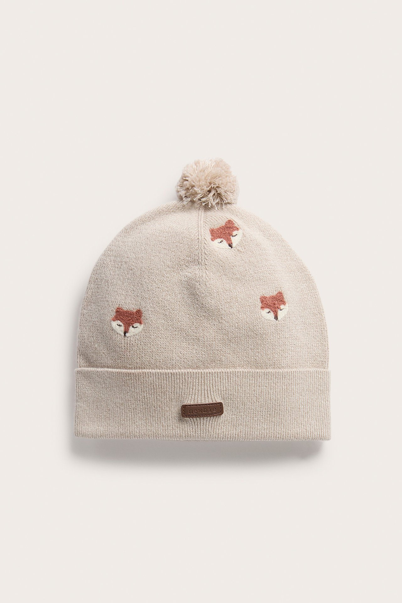 Vorderansicht einer beigen Strickmütze mit Bommel, Fuchsstickereien und einem kleinen braunen Etikett.