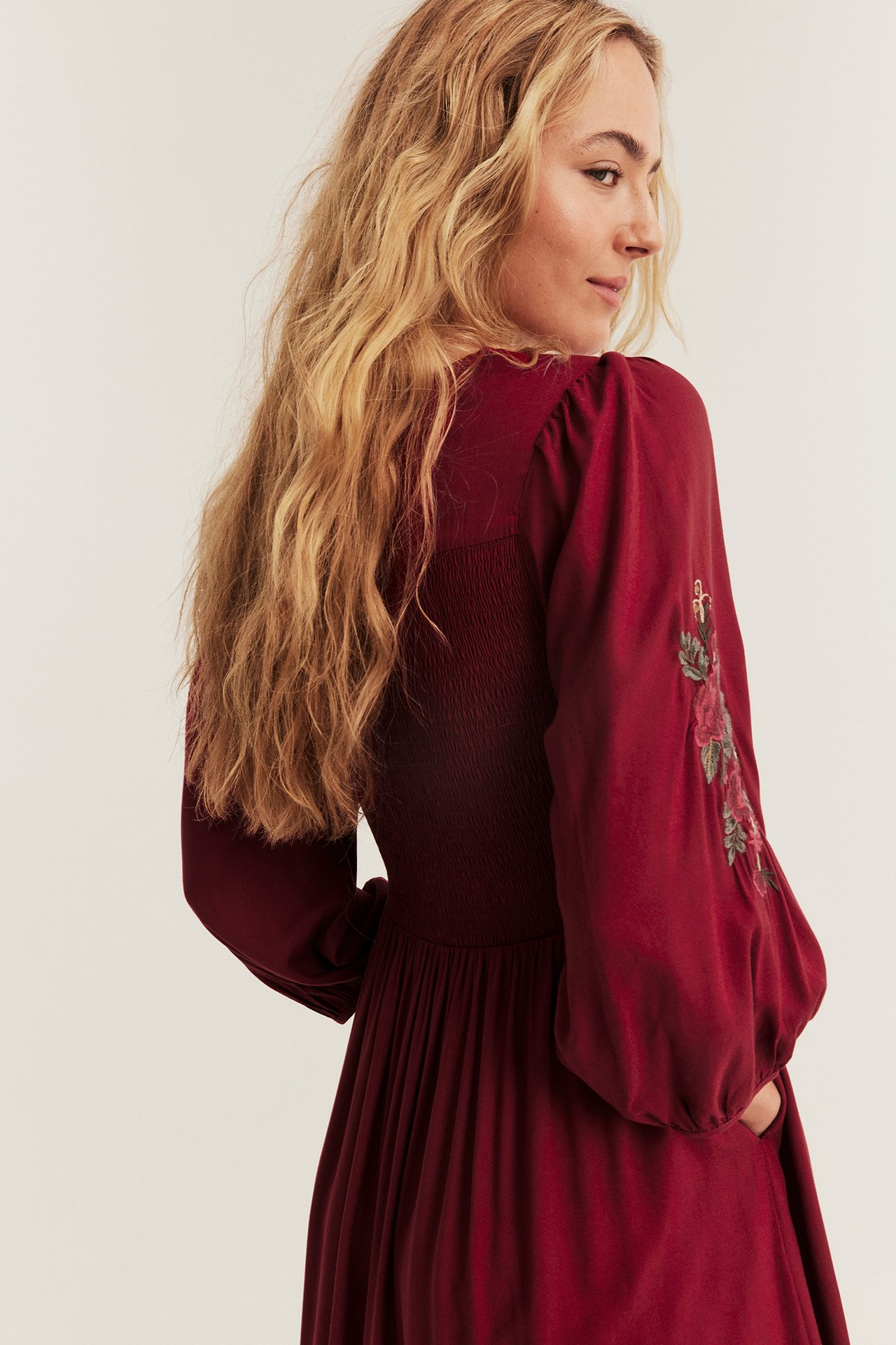 Side view of woman in red long-sleeved dress with smocked bodice, embroidered flowers.
