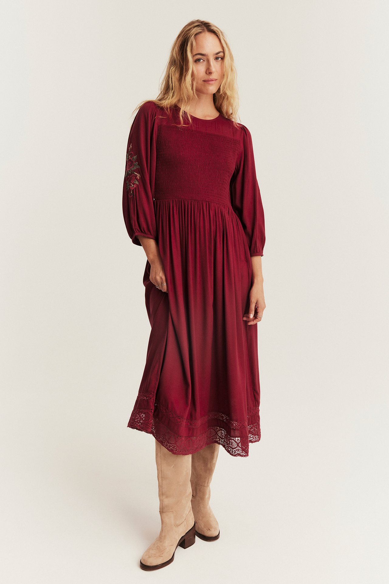 Woman wearing a red smocked midi dress with embroidered sleeves and beige boots.