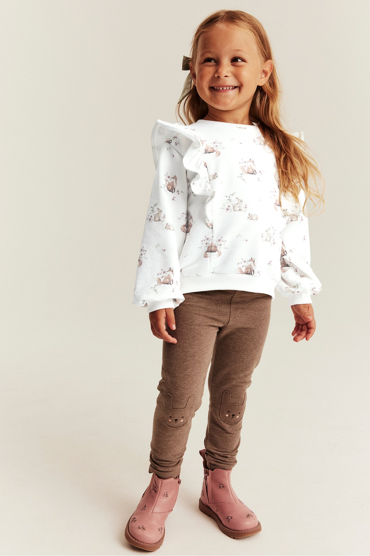 Front view of a kid wearing a white animal print ruffled sweatshirt, brown bunny knee leggings, and pink embroidered boots.