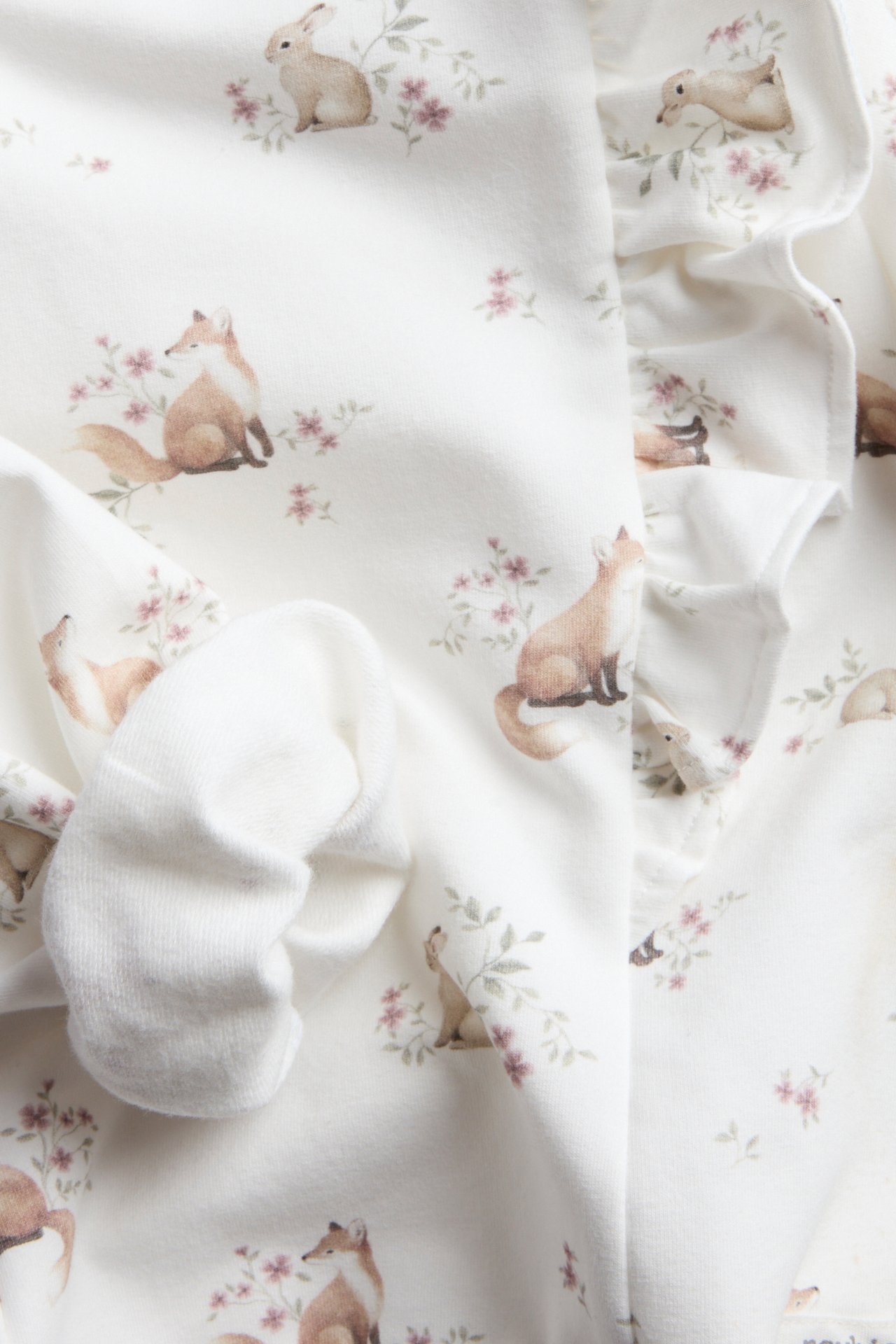 Nahaufnahme von weißem Babykleidungsstück mit Fuchs- und Hasenprint und Rüsche, neben einem weißen Babysocken.