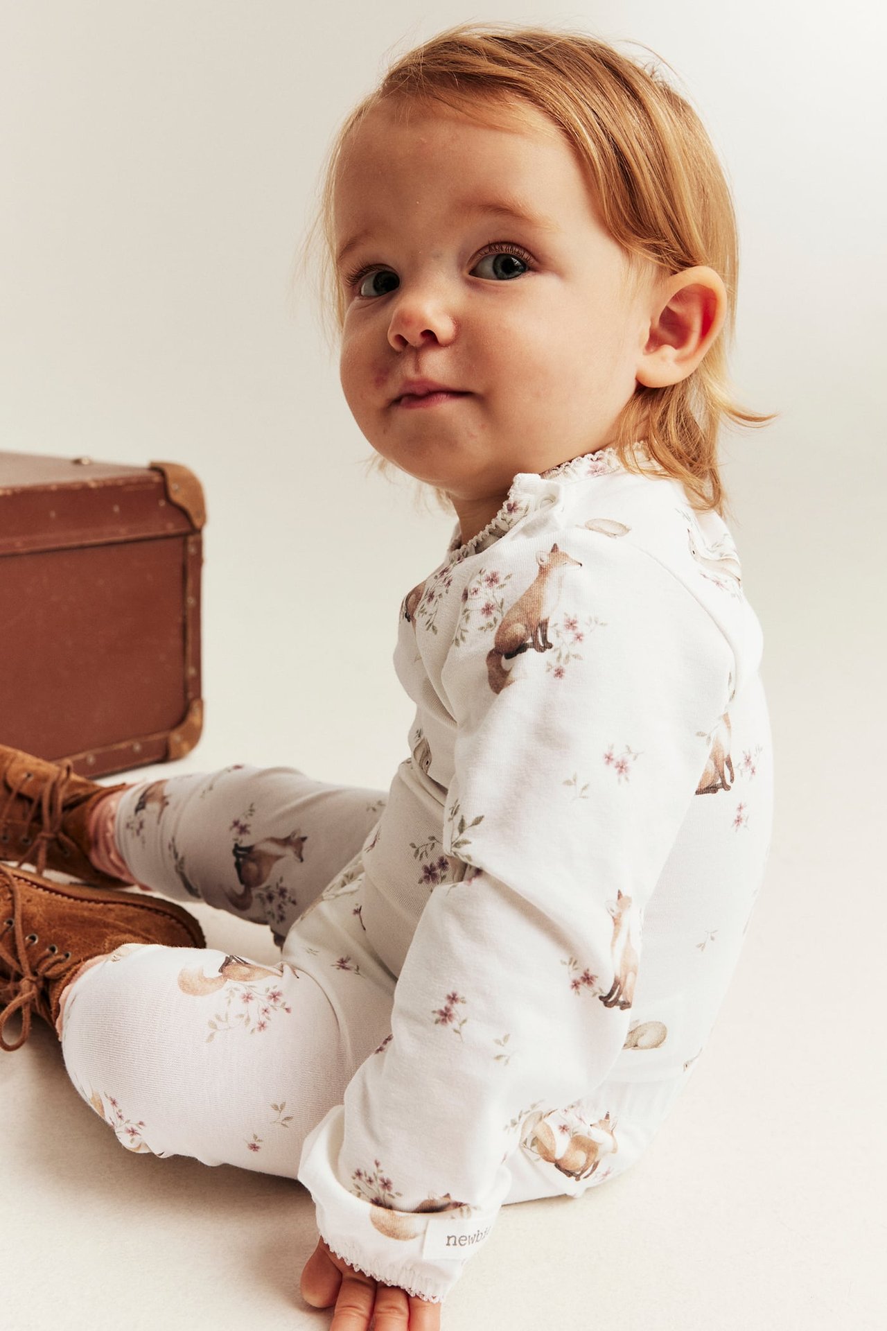 Baby von vorne in weißem, langärmeligem Overall mit Fuchs- und Blumenmuster und braunen Schnürstiefeln.