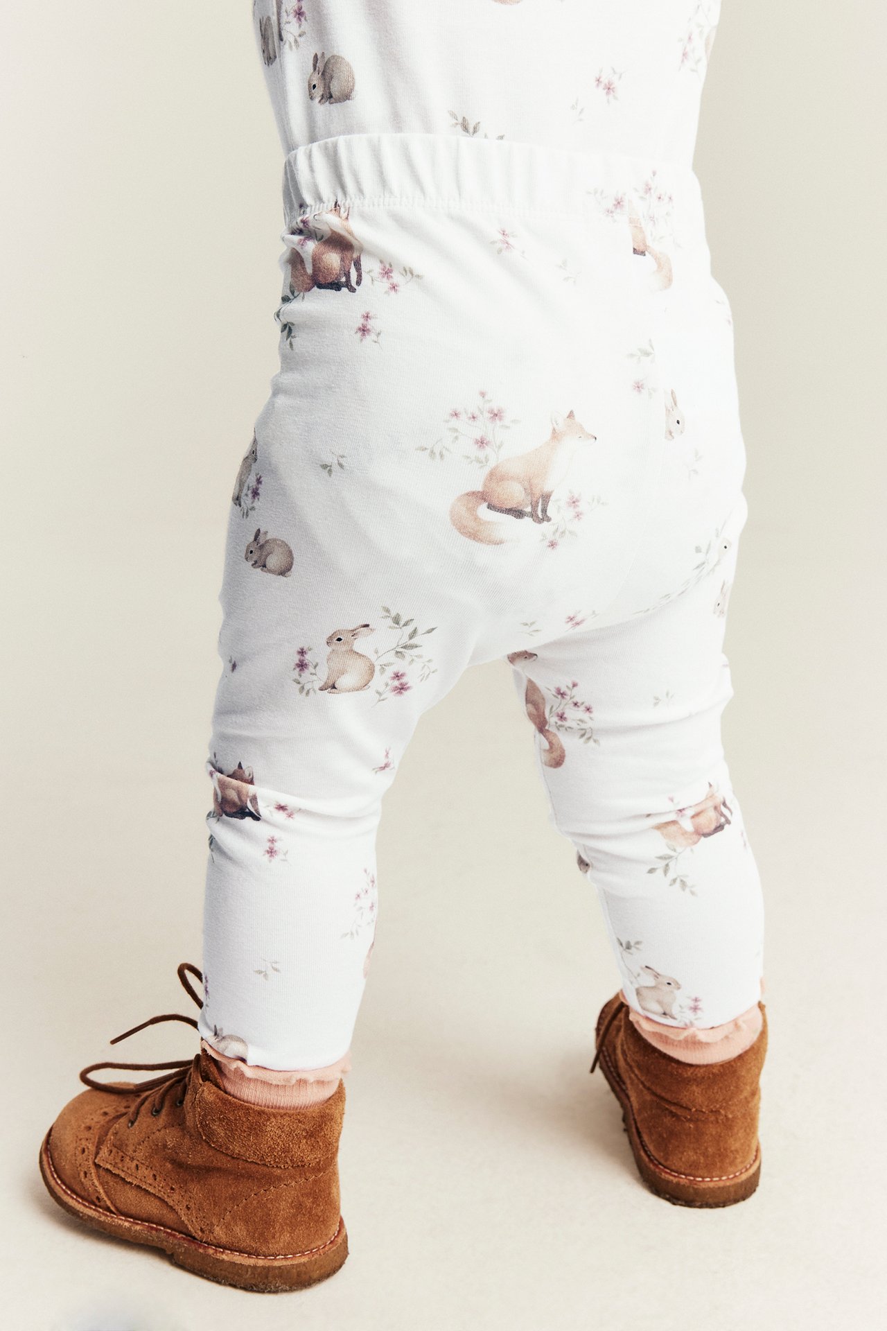 Rückansicht: Baby in weißen Leggings mit Tiermuster und braunen Schnürstiefeln.