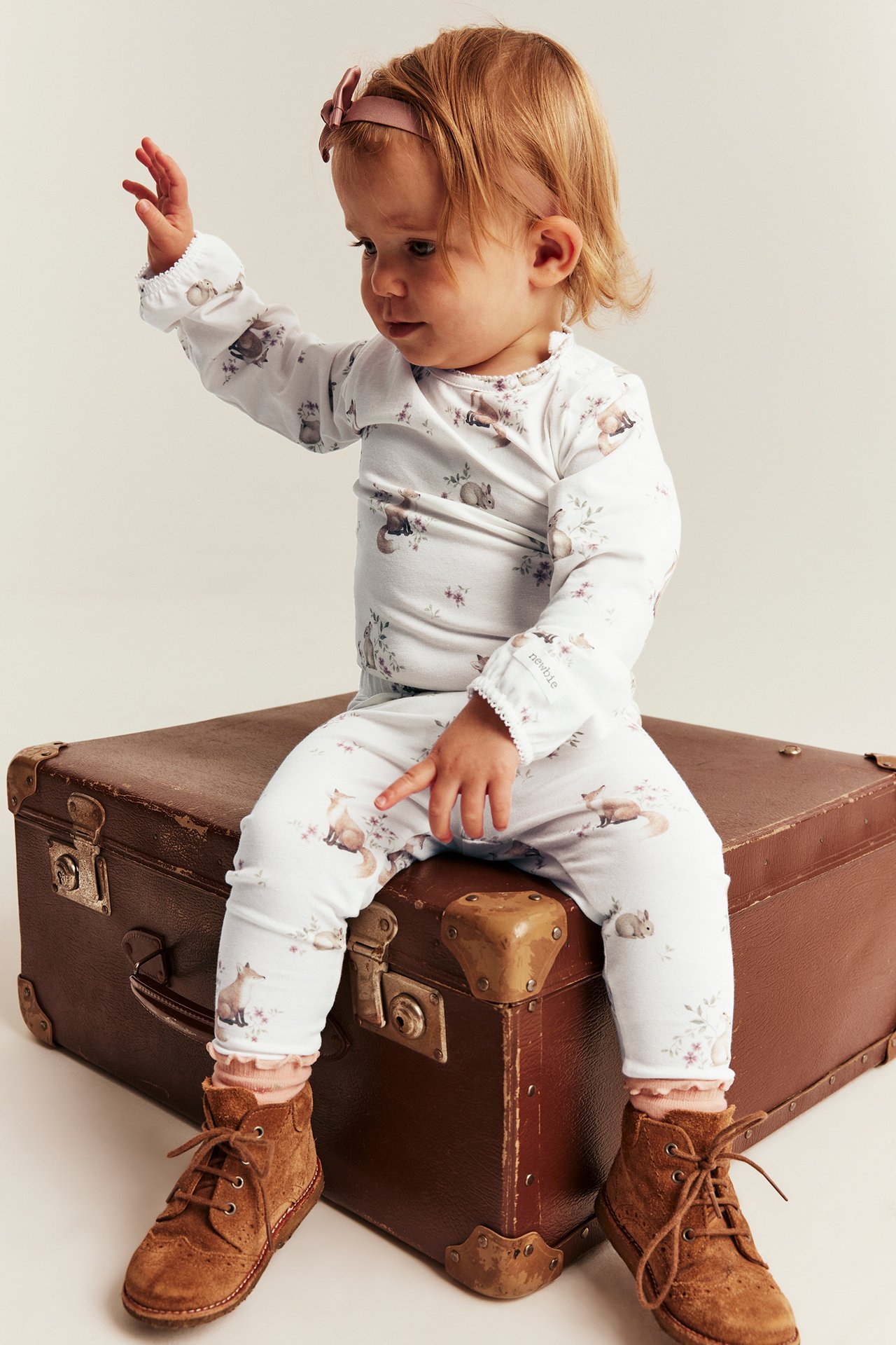 Vorderansicht: Baby in weißem Tiermuster-Set, braunen Stiefeln und rosa Haarband.