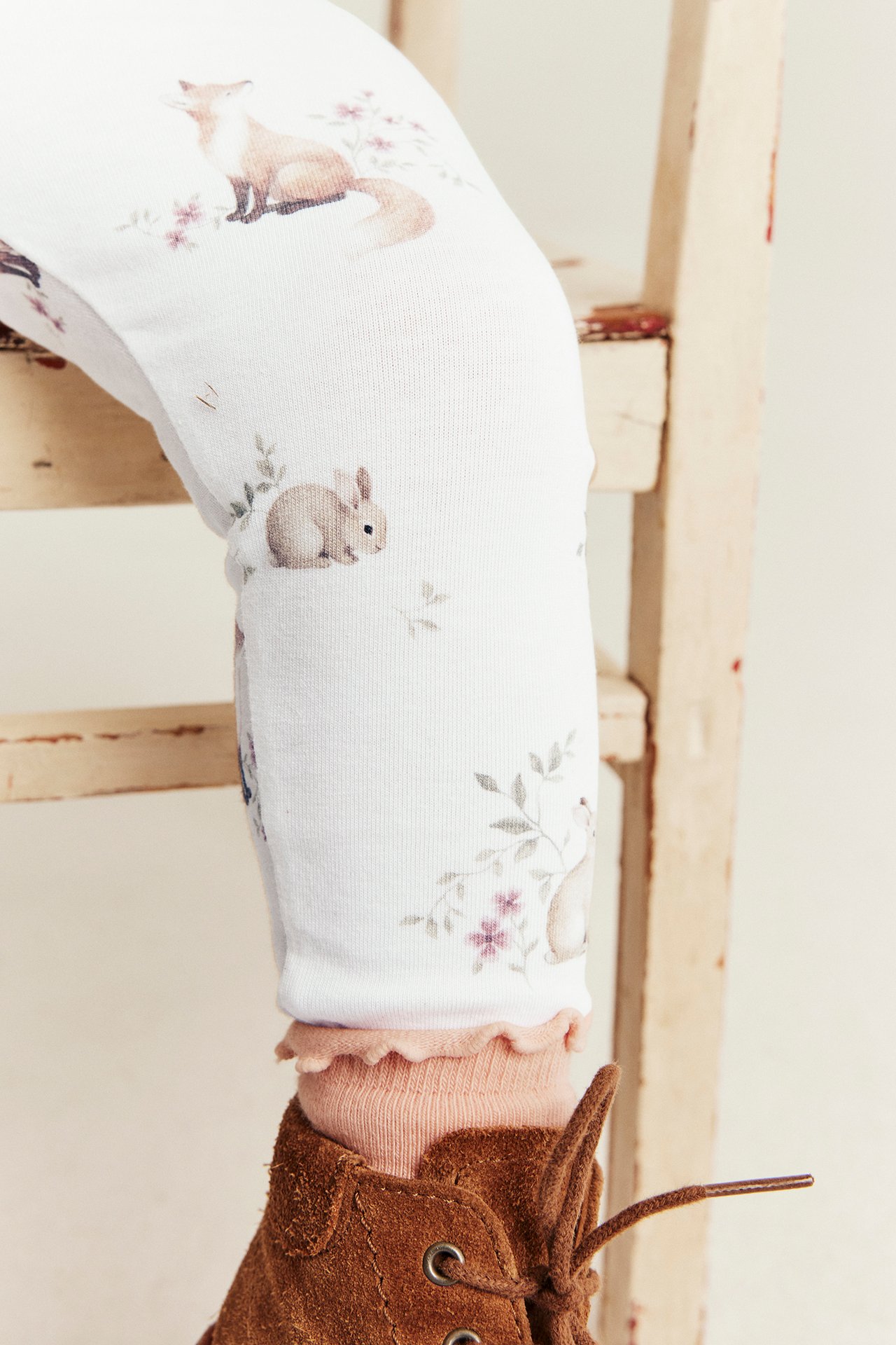 Nahaufnahme eines Kinderbeins in weißen bedruckten Leggings, rosa Socken und braunen Wildleder-Schnürstiefeln.