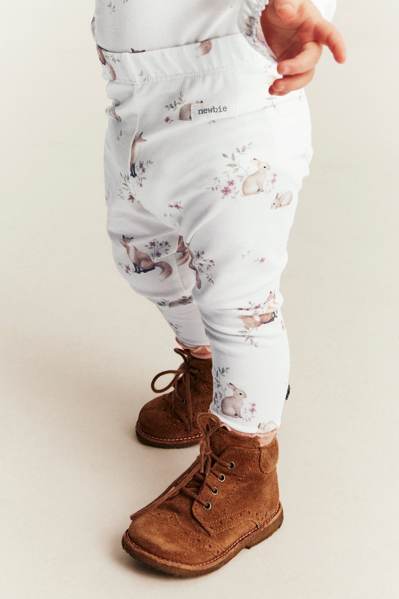 Vorderansicht eines Babys in weißen Leggings mit Fuchs- und Hasenmotiv und braunen Schnürstiefeln.