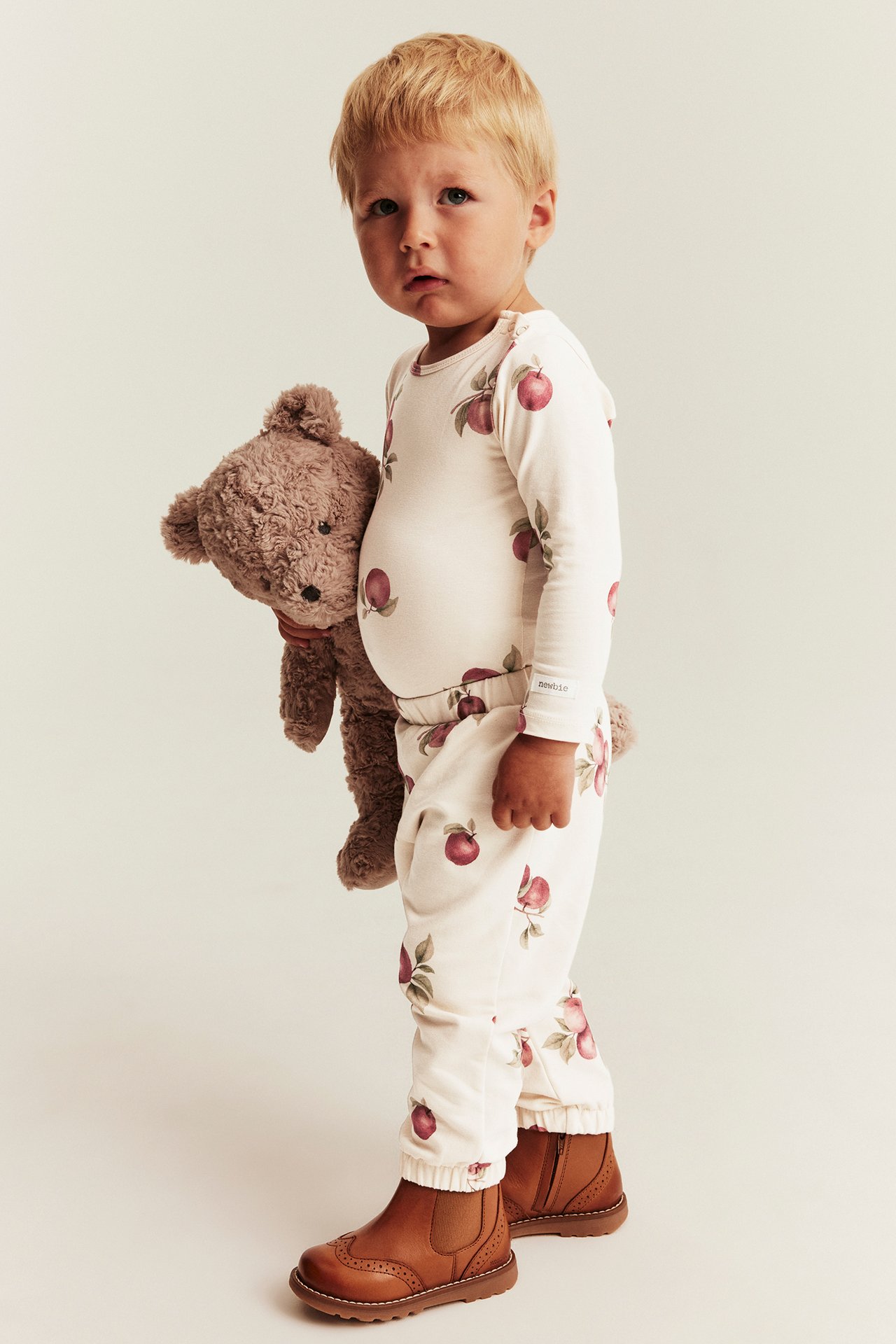 Side view: Kid in cream apple print pajama set and brown boots, holding a brown teddy bear.