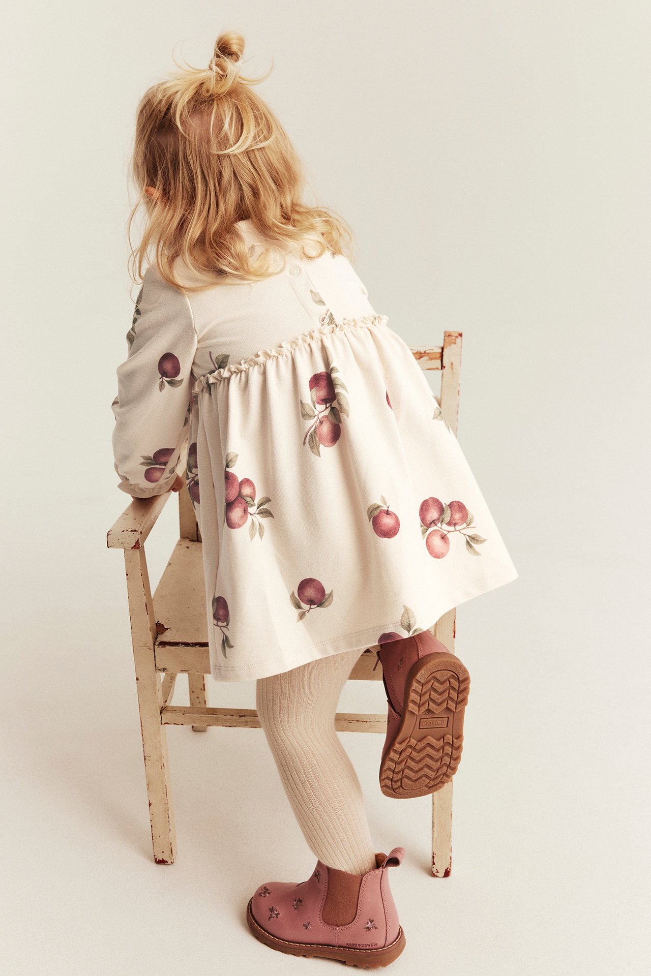Kid's back view: beige long-sleeve apple print dress, white tights, pink embroidered boots.