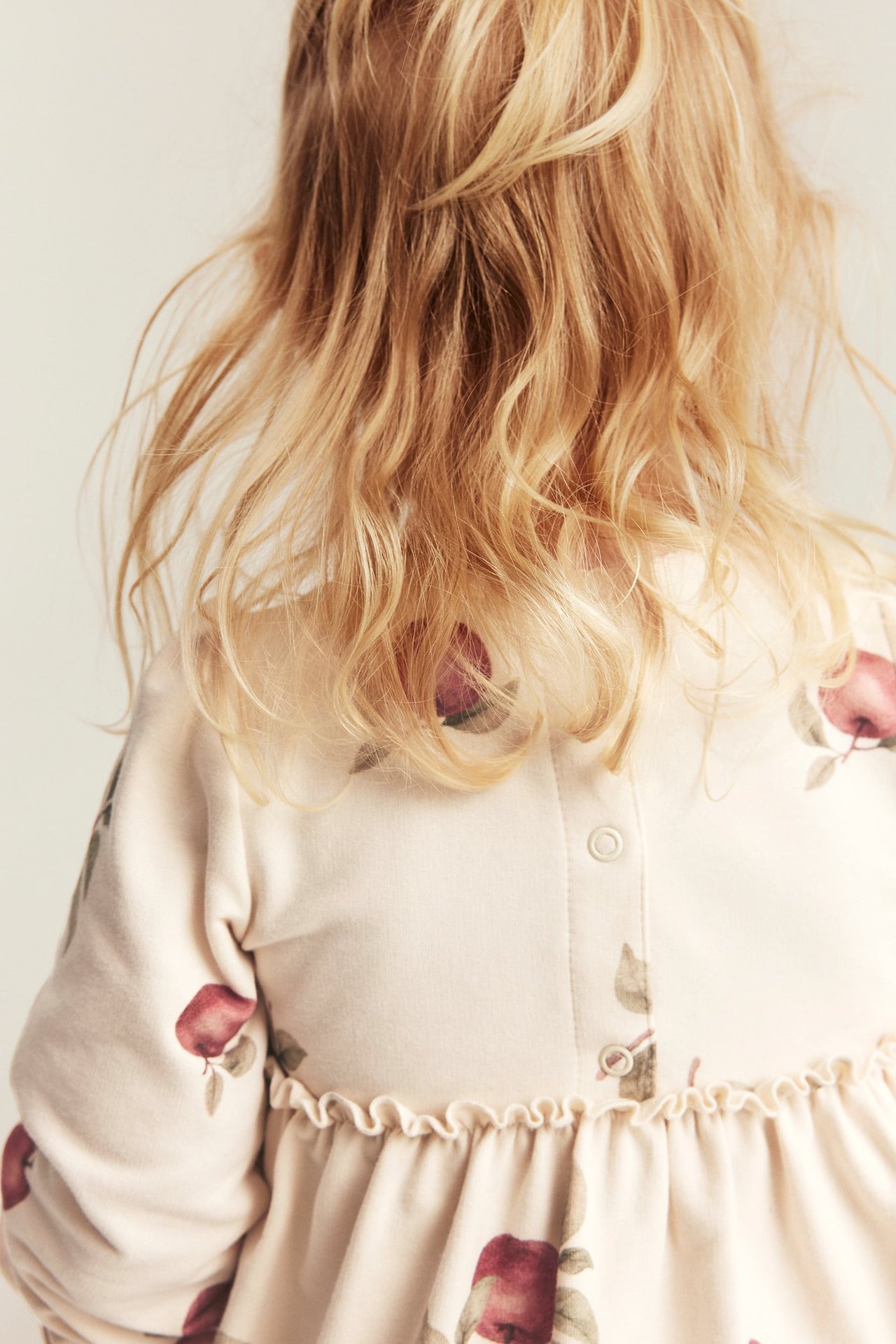 Close-up back view of a kid in a beige long-sleeve dress with red apple print and back buttons.