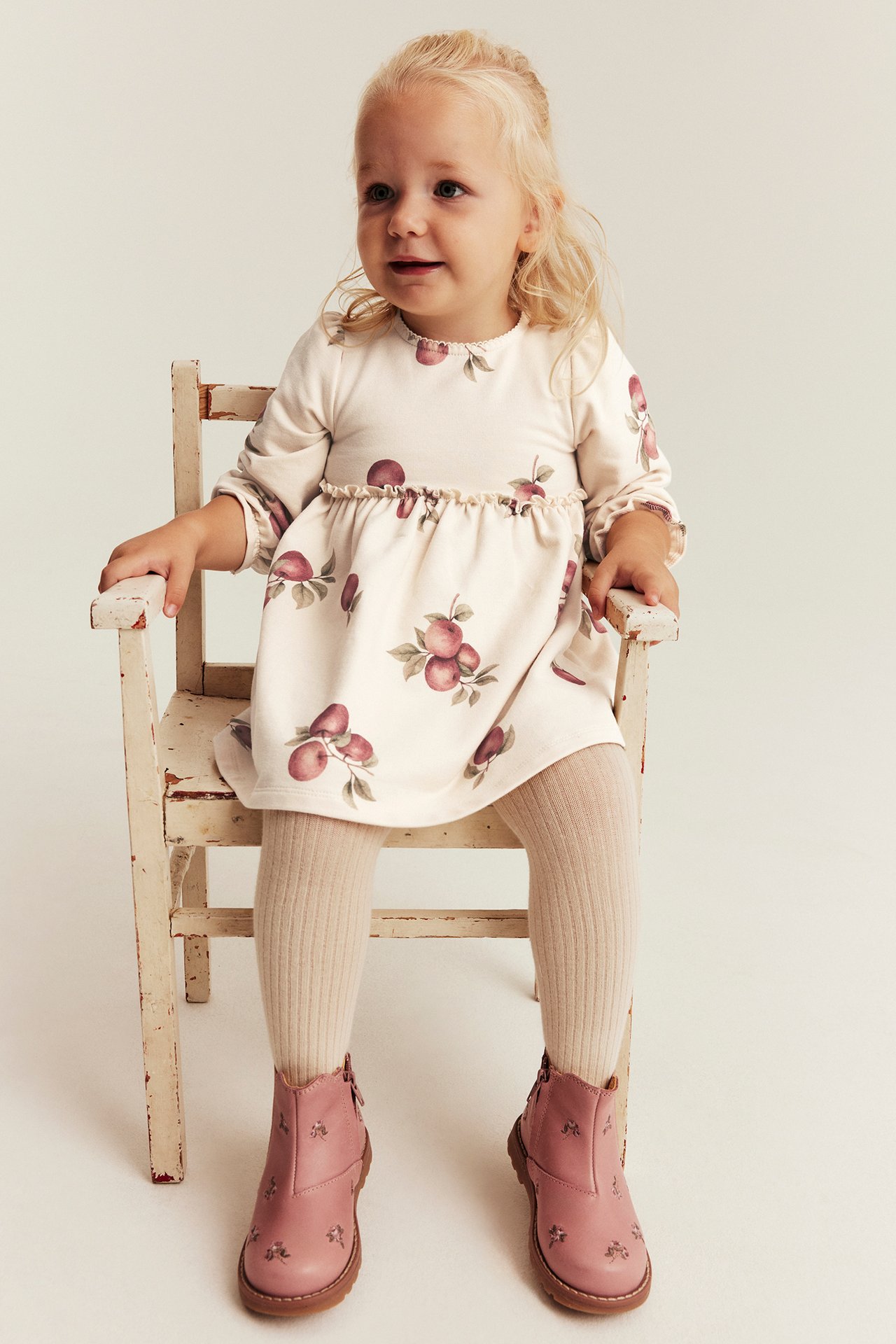Front view of a baby girl in a beige apple print dress, ribbed beige tights, and pink embroidered ankle boots.
