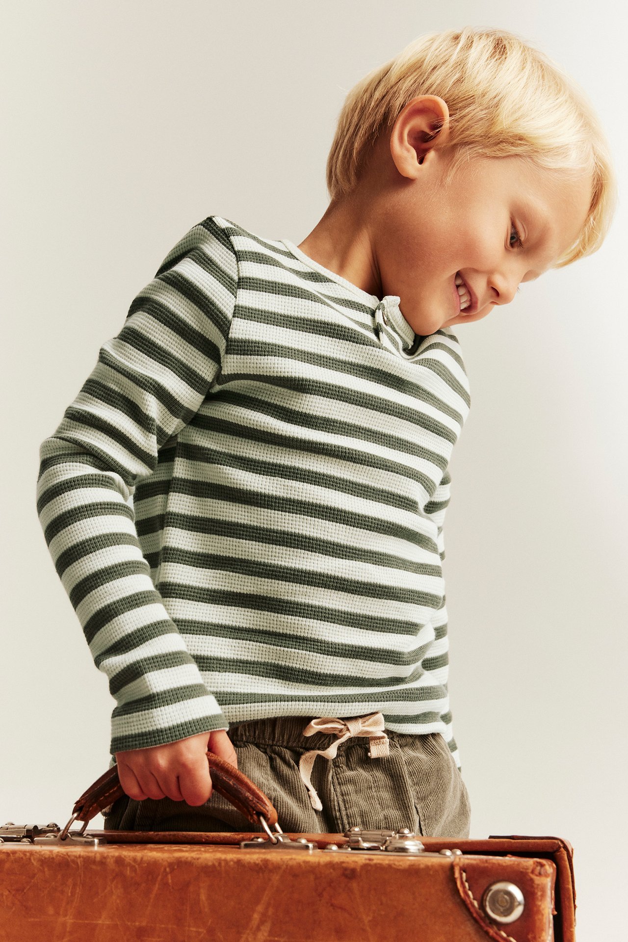 Grün-weiß gestreiftes Waffelstrick-Langarmshirt für Kinder, mit Khaki-Kordhosen, einen braunen Koffer haltend.