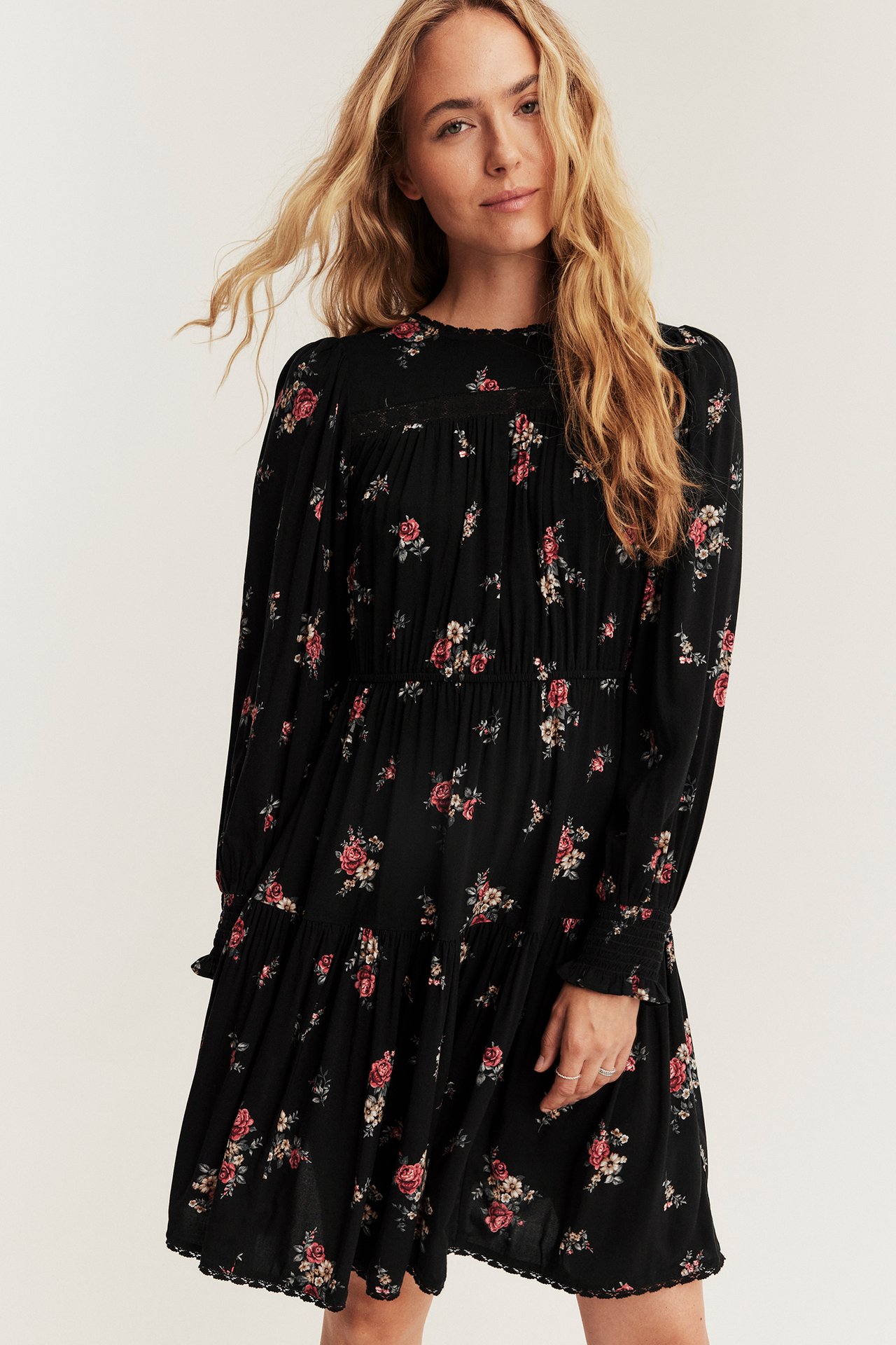 Vorderansicht: weibliches Model trägt schwarzes geblümtes Stufen-Minikleid mit Smokbündchen, Spitze, Ringen.