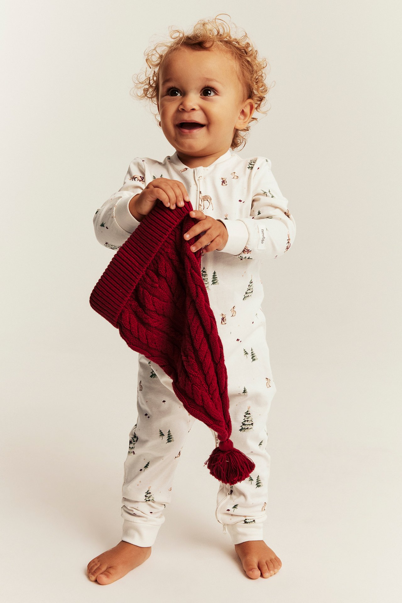 Front view: Baby in white long-sleeved forest print pajama, holding a red cable-knit hat.