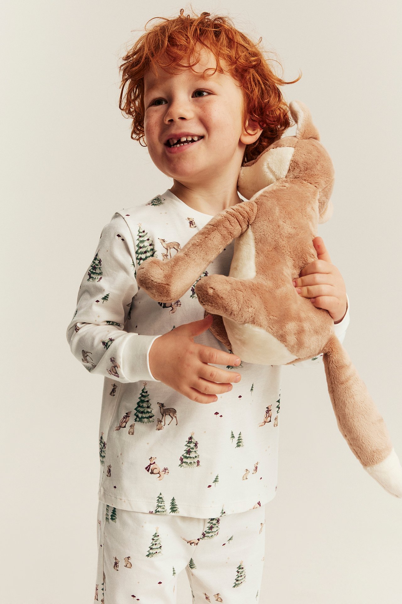 Front view of a kid in white pajamas with a forest print, holding a brown plush toy.
