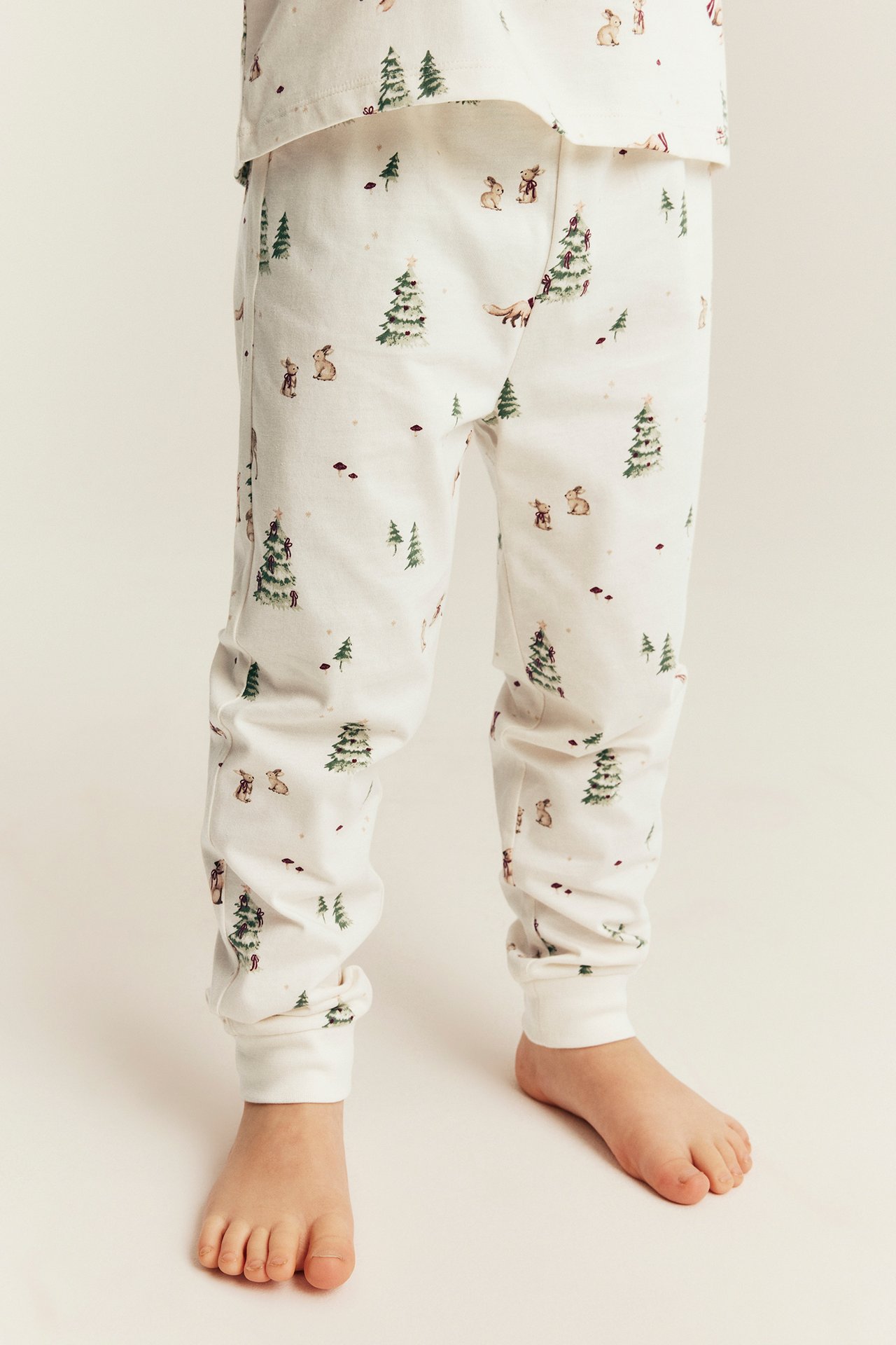 Front view of a kid wearing white pajama pants with woodland animal and tree print and a matching top.